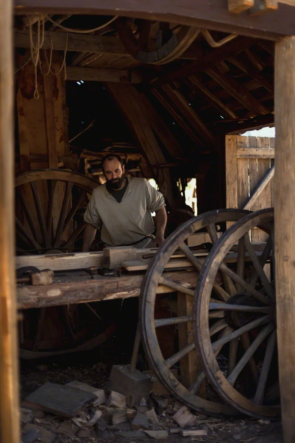 A man with a beard in a light-colored shirt inside a wooden barn with large wagon wheels and tools, looking thoughtfully at the camera.