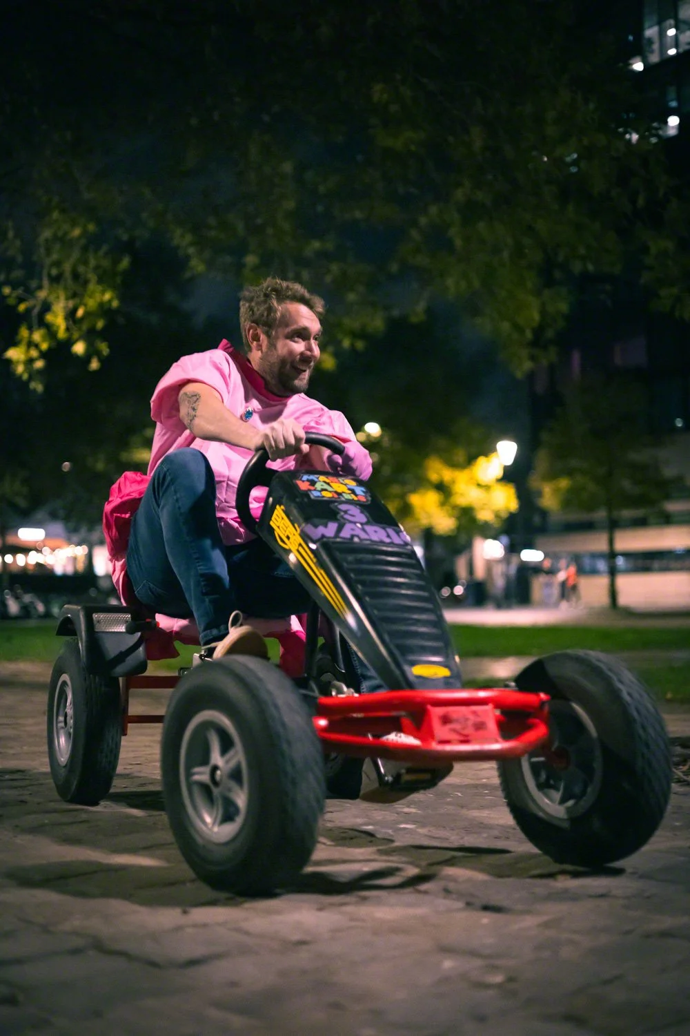 A man with a pink shirt driving a small pedal-powered go-kart at night in an outdoor urban setting.