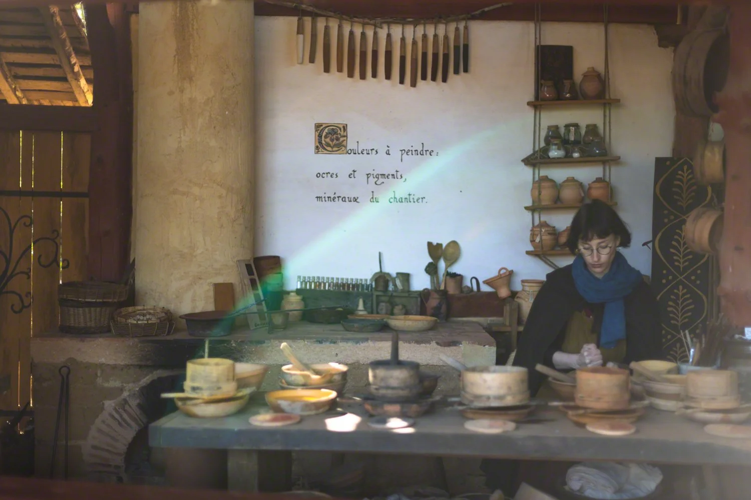 A woman in historical costume working at a pottery table in a rustic workshop with pottery shelves and writing on the wall