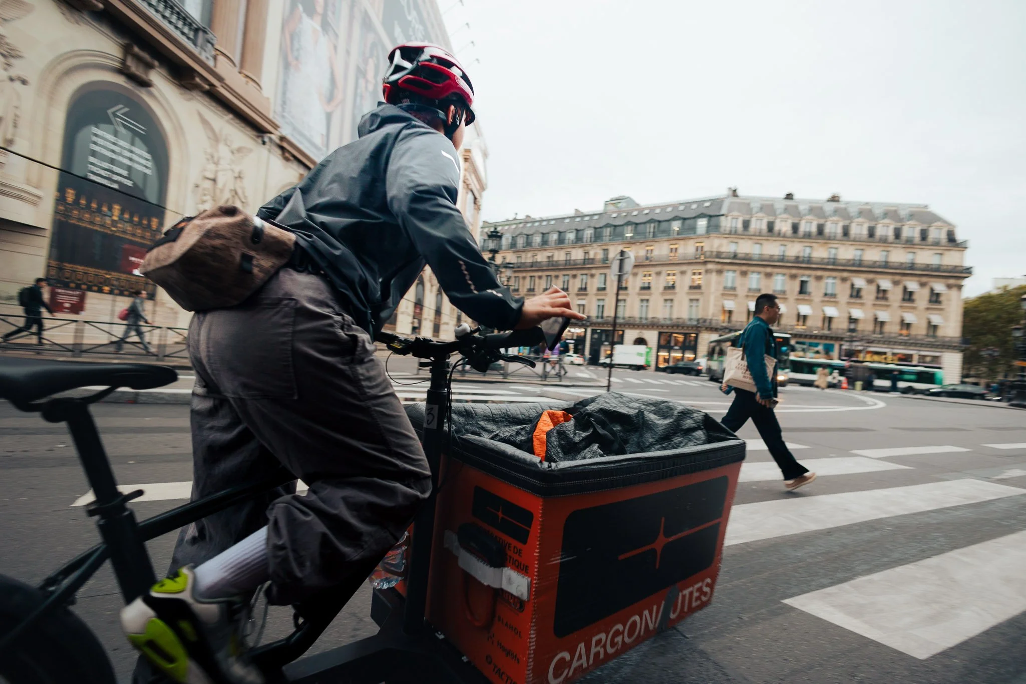 A cyclist riding a delivery bike across a city street, with pedestrians walking and historic buildings in the background.