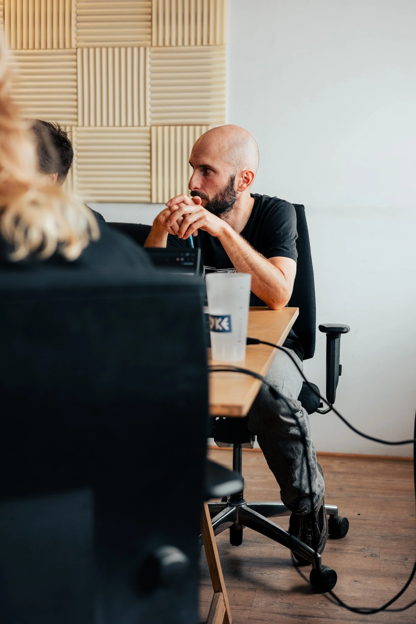 A bald man with a beard sitting at a conference table, resting his chin on his hands and looking to the side, in a room with acoustic wall panels.