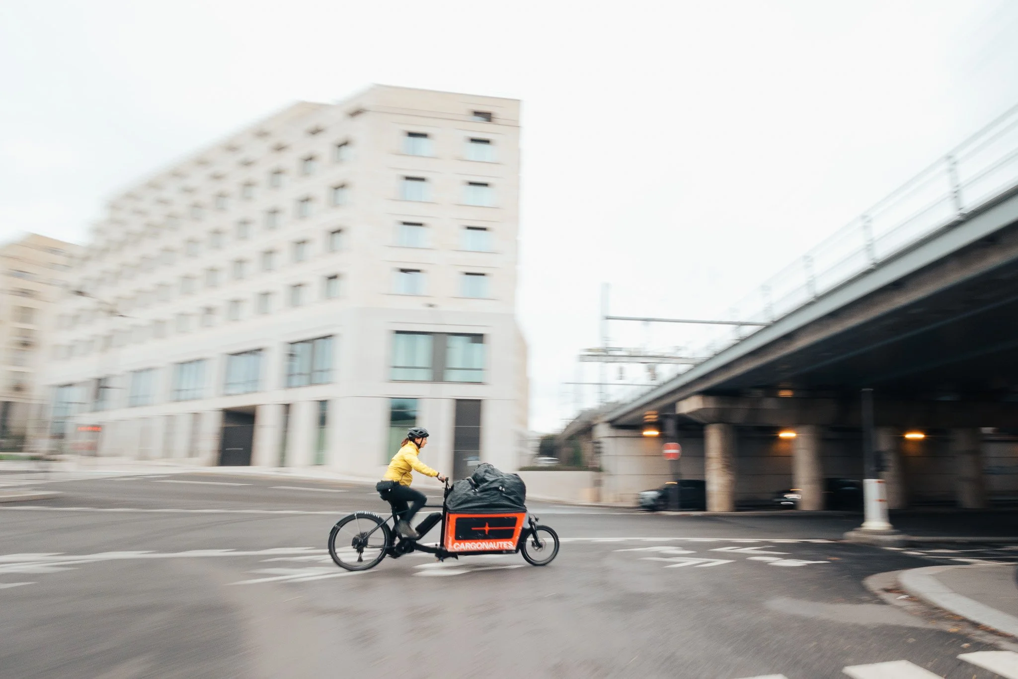 A cyclist riding a cargo bike across a city street, with a large building and an elevated road in the background.