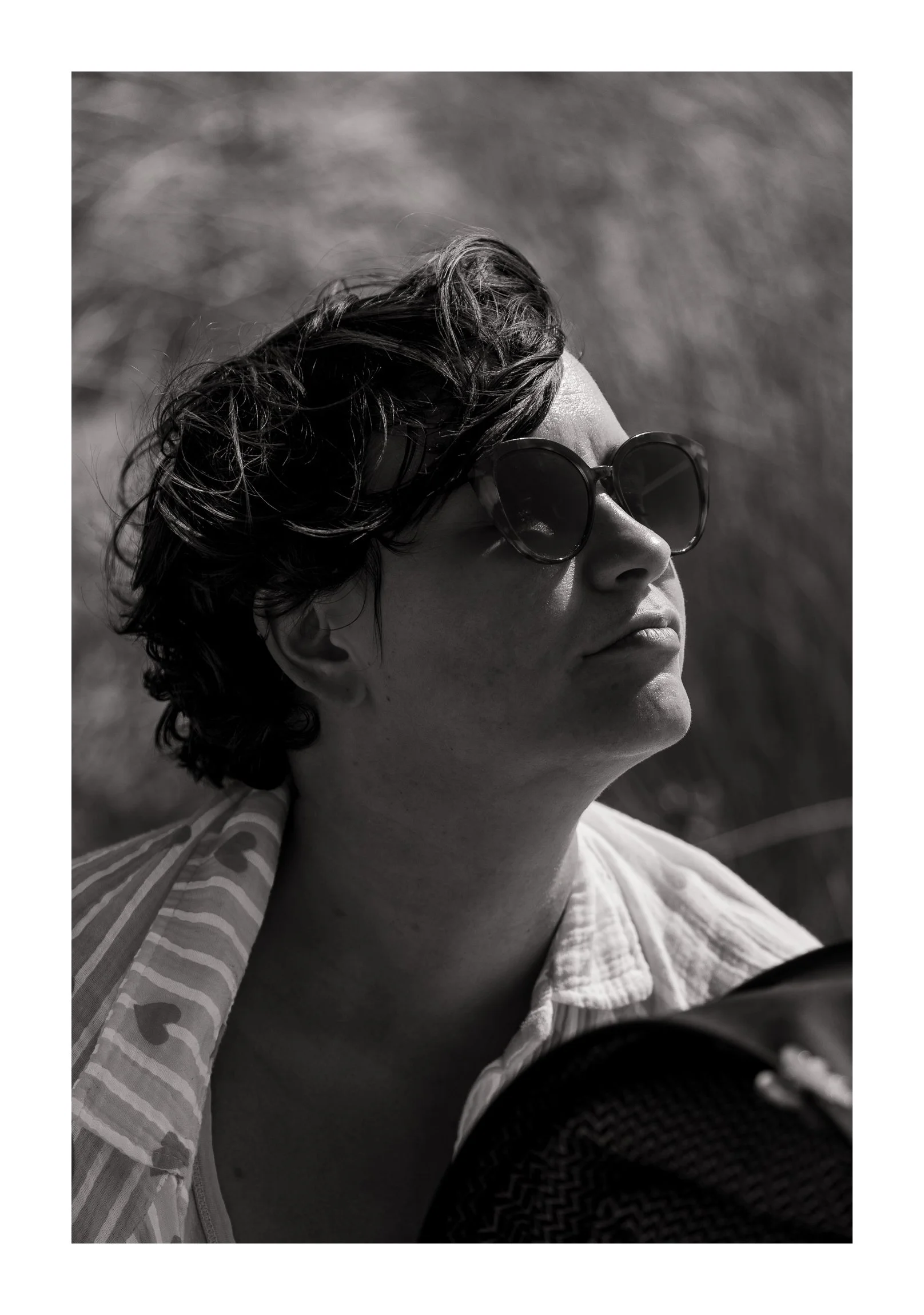 A woman with short wavy hair wearing large sunglasses and a striped shirt, looking up and to the right outdoors in black and white.