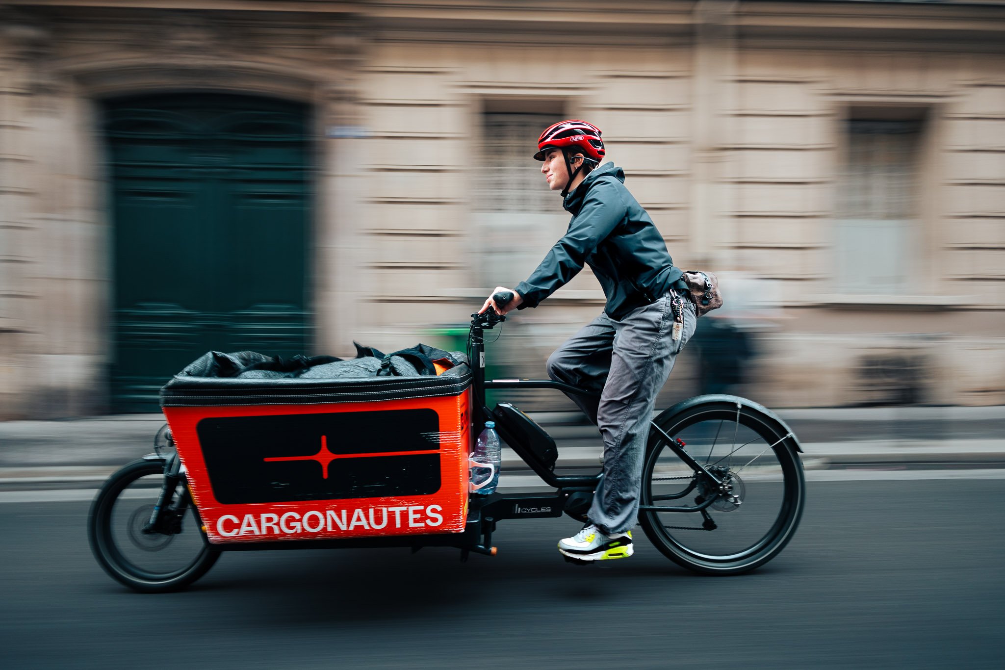 A man riding a cargo e-bike with a large red and black cargo box labeled 'CARGONAUTES' down an urban street, wearing a red helmet and gray clothes.