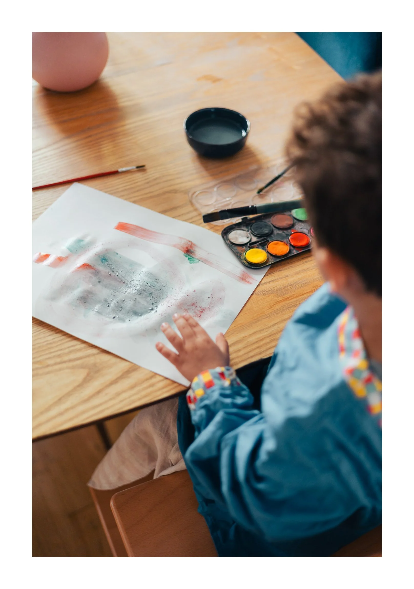 Child wearing a blue jacket and beige pants is painting with watercolors on a white sheet of paper at a wooden table. Art supplies, including a paintbrush, paint palette with various colors, and a small water container, are on the table. A pink objec