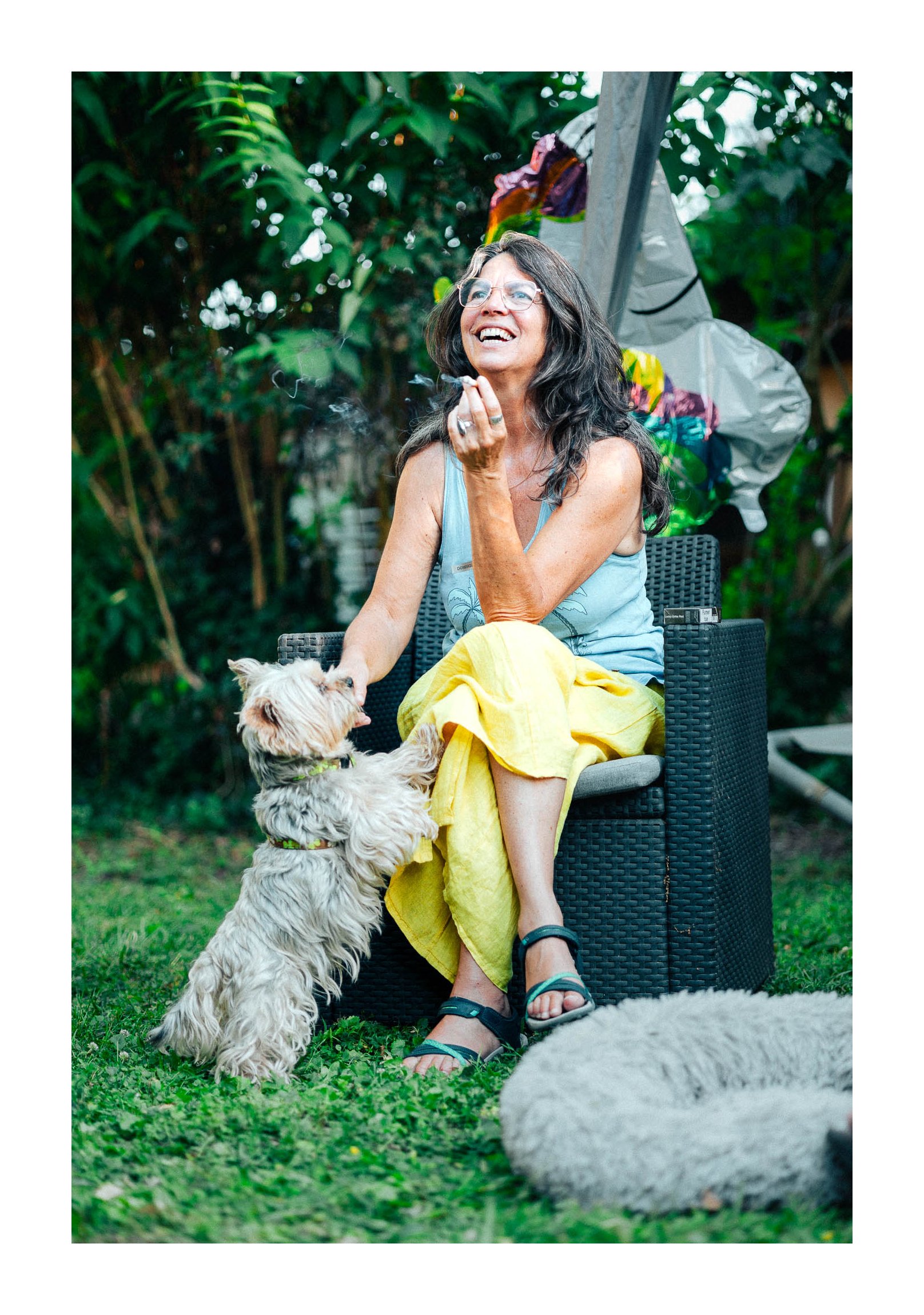 A woman with long dark hair, glasses, and a light blue tank top sitting outdoors on a black chair, smiling and holding a cigarette while playing with a fluffy light-colored dog that is standing on its hind legs. There is another dog lying on the gras