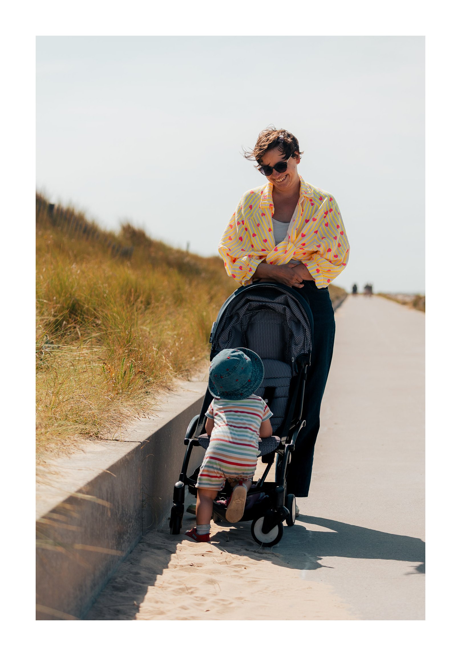 A woman with short dark hair and sunglasses smiling at a young child in a stroller on a sandy path during a sunny day, with grassy dunes to the side.