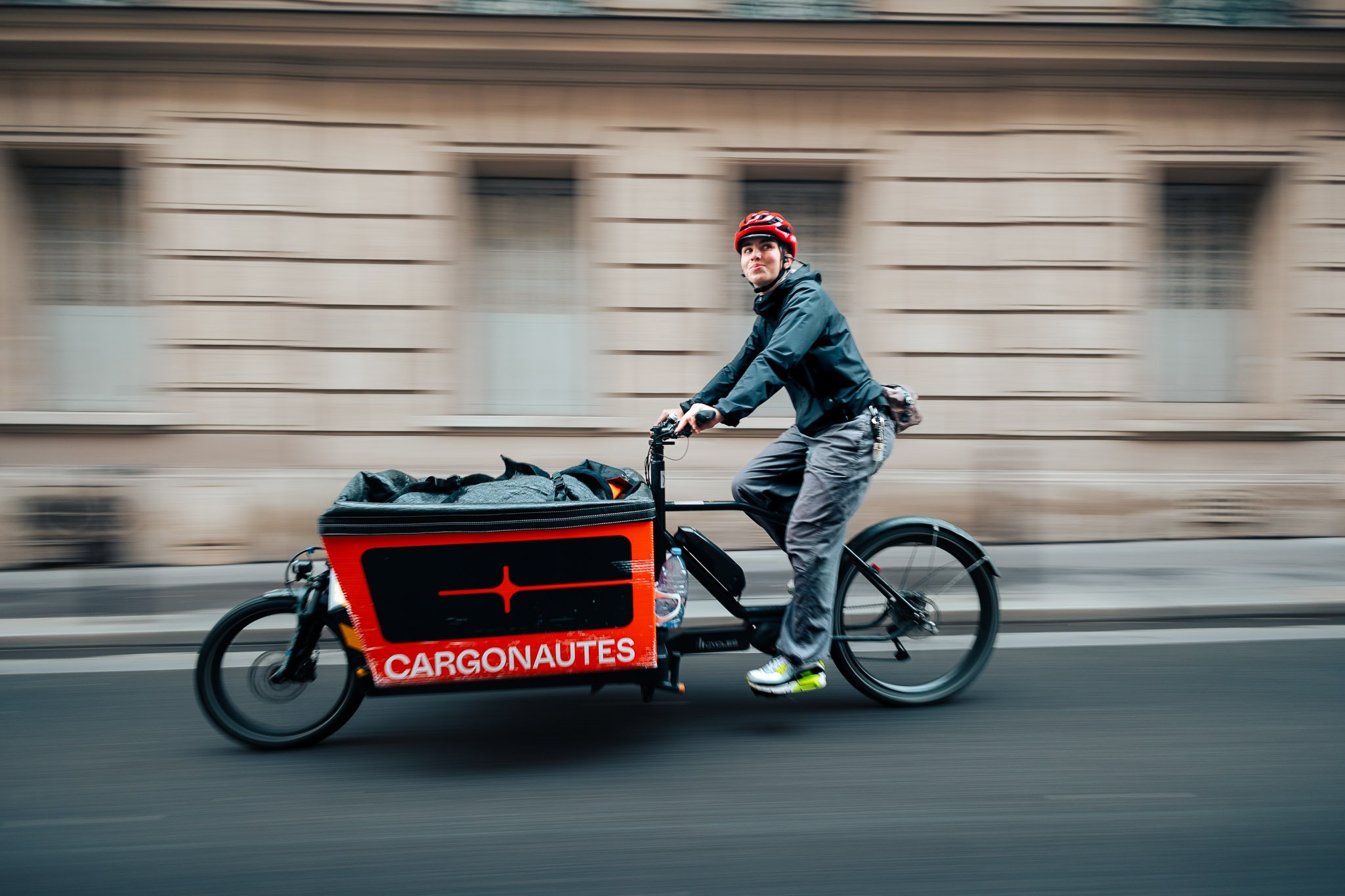 Person riding a cargo bicycle with a sign that says 'CARGONAUTES', blurred background, wearing a helmet and dark clothing.