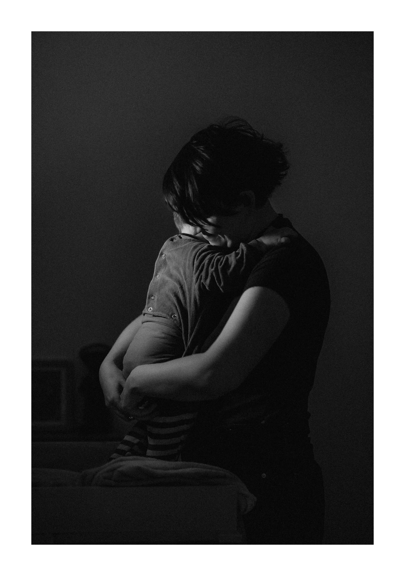 A black-and-white photo of a person hugging a child, with both facing each other and their faces close together. The person has dark hair and is wearing a dark shirt, while the child has short hair, striped pants, and a long-sleeved shirt. The backgr