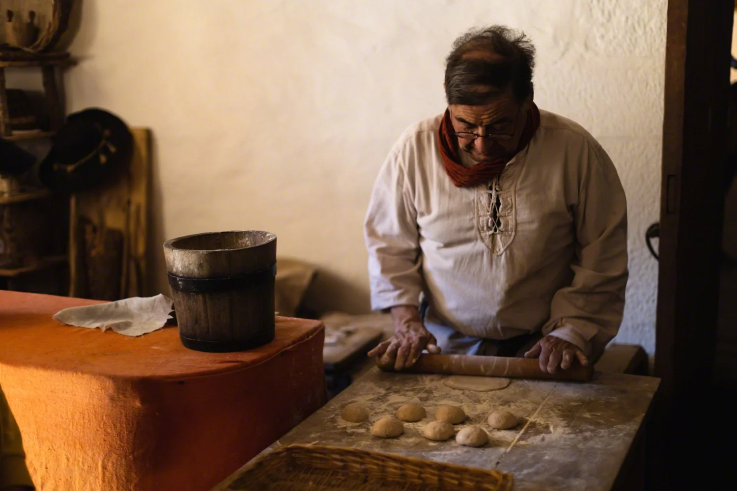An elderly man with glasses and a red scarf around his neck is rolling out dough on a wooden table, with several round pieces of dough in front of him. A wooden container and a cloth are on the table, and the background features a beige wall and a wo