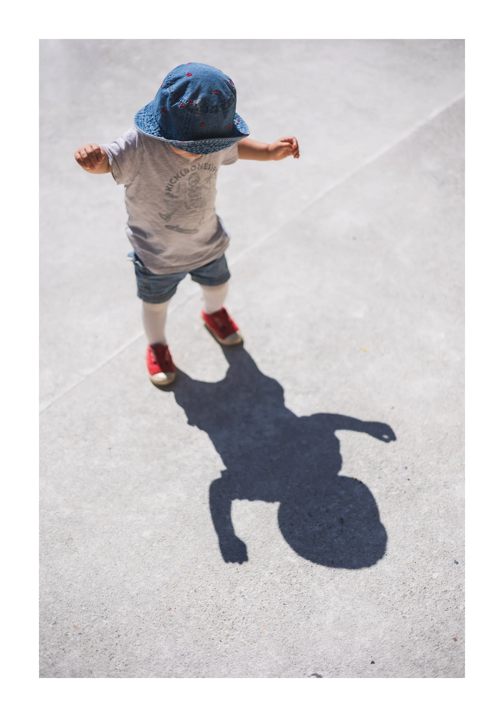 A young child wearing a blue hat, grey T-shirt, shorts, white socks, and red shoes stands on a concrete surface, creating a shadow that resembles a dog.