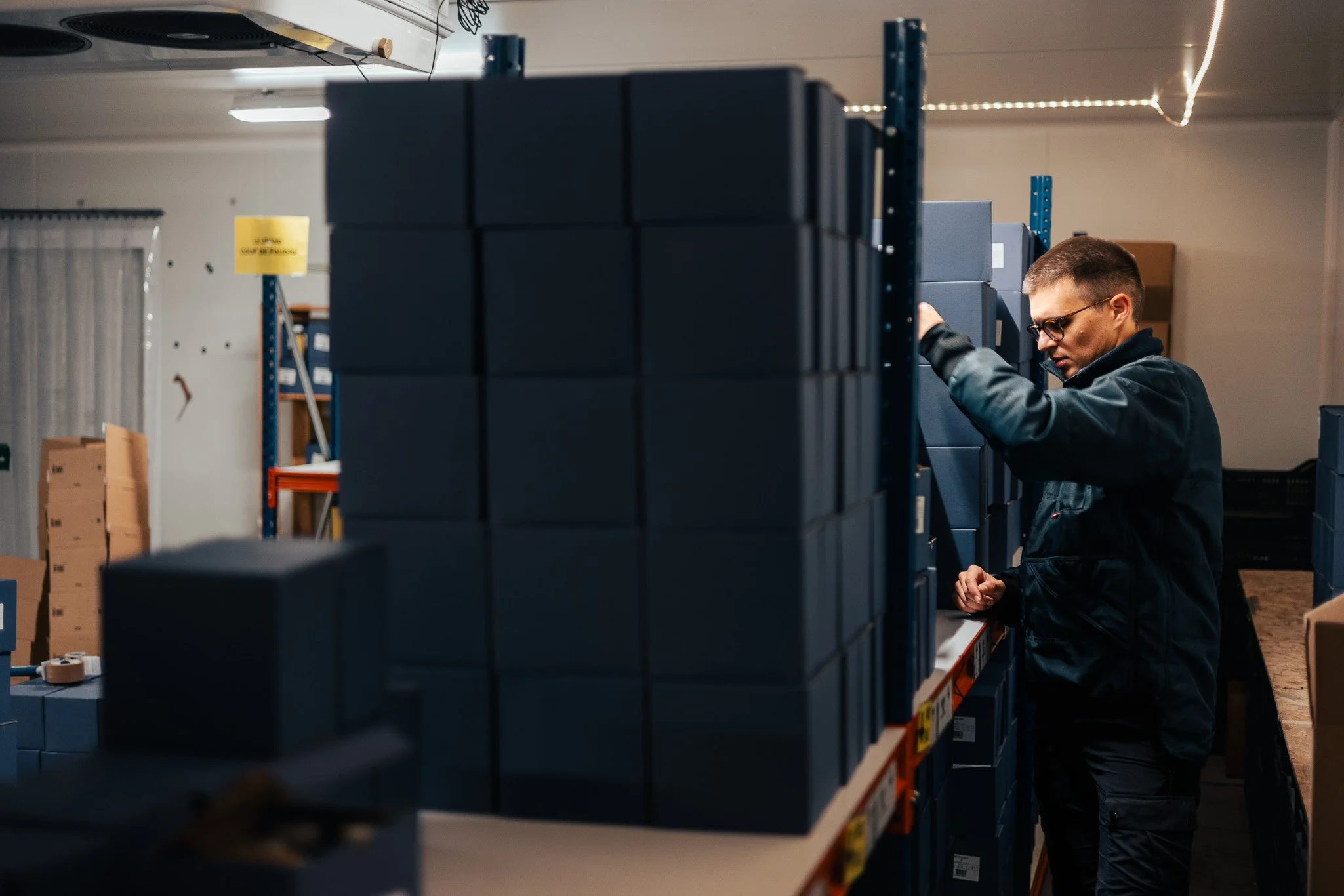 A man in black jacket and glasses standing in a warehouse, inspecting or organizing dark gray boxes on metal shelves.