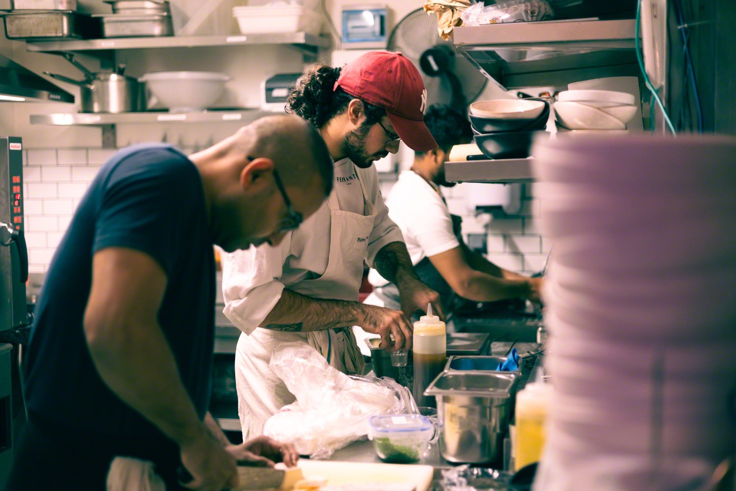 Three men preparing food in a commercial kitchen, one cutting ingredients, another seasoning, and a third working in the background.