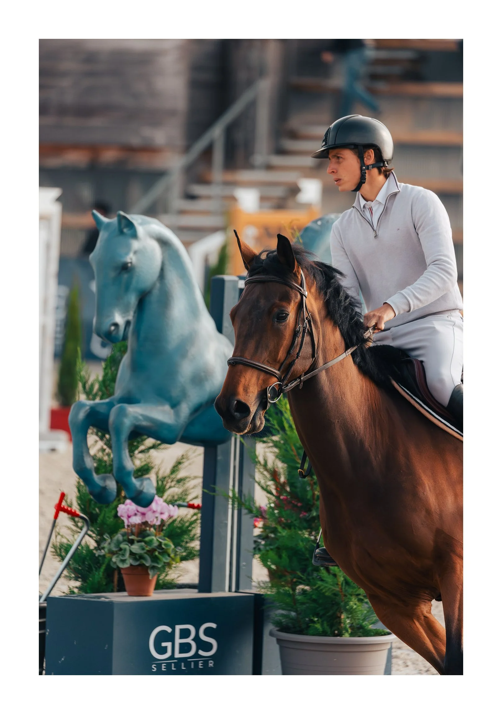 A woman wearing riding gear and a helmet riding a brown horse at an equestrian event, approaching a horse sculpture with a sign that reads 'GBS SELLER' in front of plants and fencing.