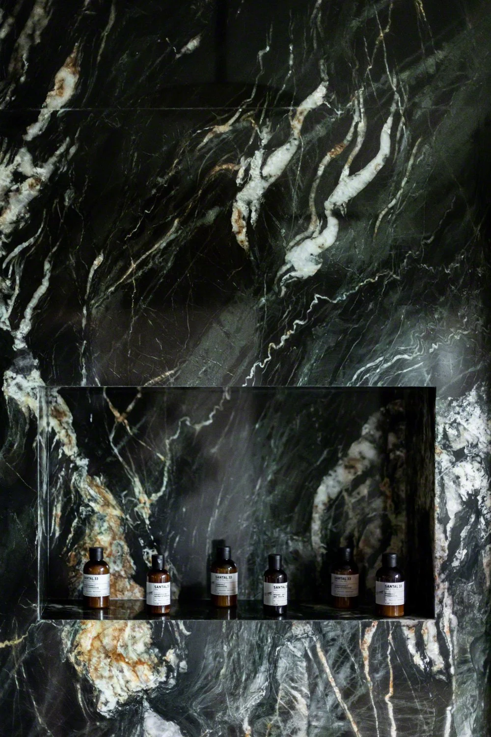 A dark marble wall with white and gold veining, featuring a recessed shelf holding six small amber bottles with white labels.