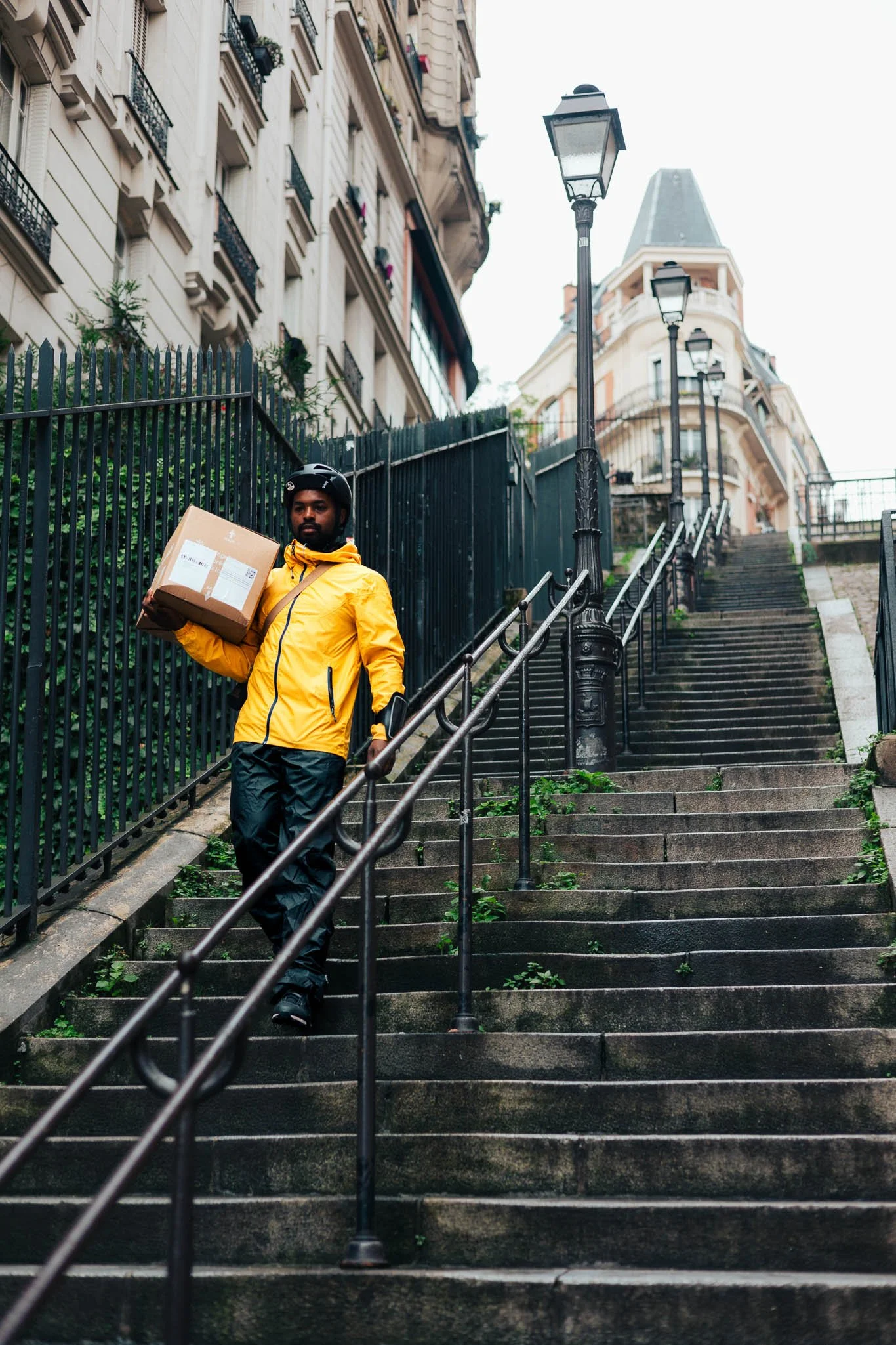 A delivery person in a yellow jacket and helmet walking down a steep outdoor staircase holding a package.