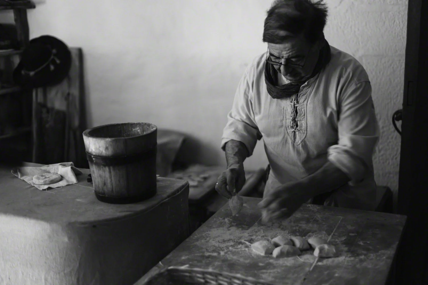 A man wearing glasses, a long-sleeve shirt, and a scarf is preparing food on a wooden table, with a pot and cloth nearby, in a domestic kitchen setting.