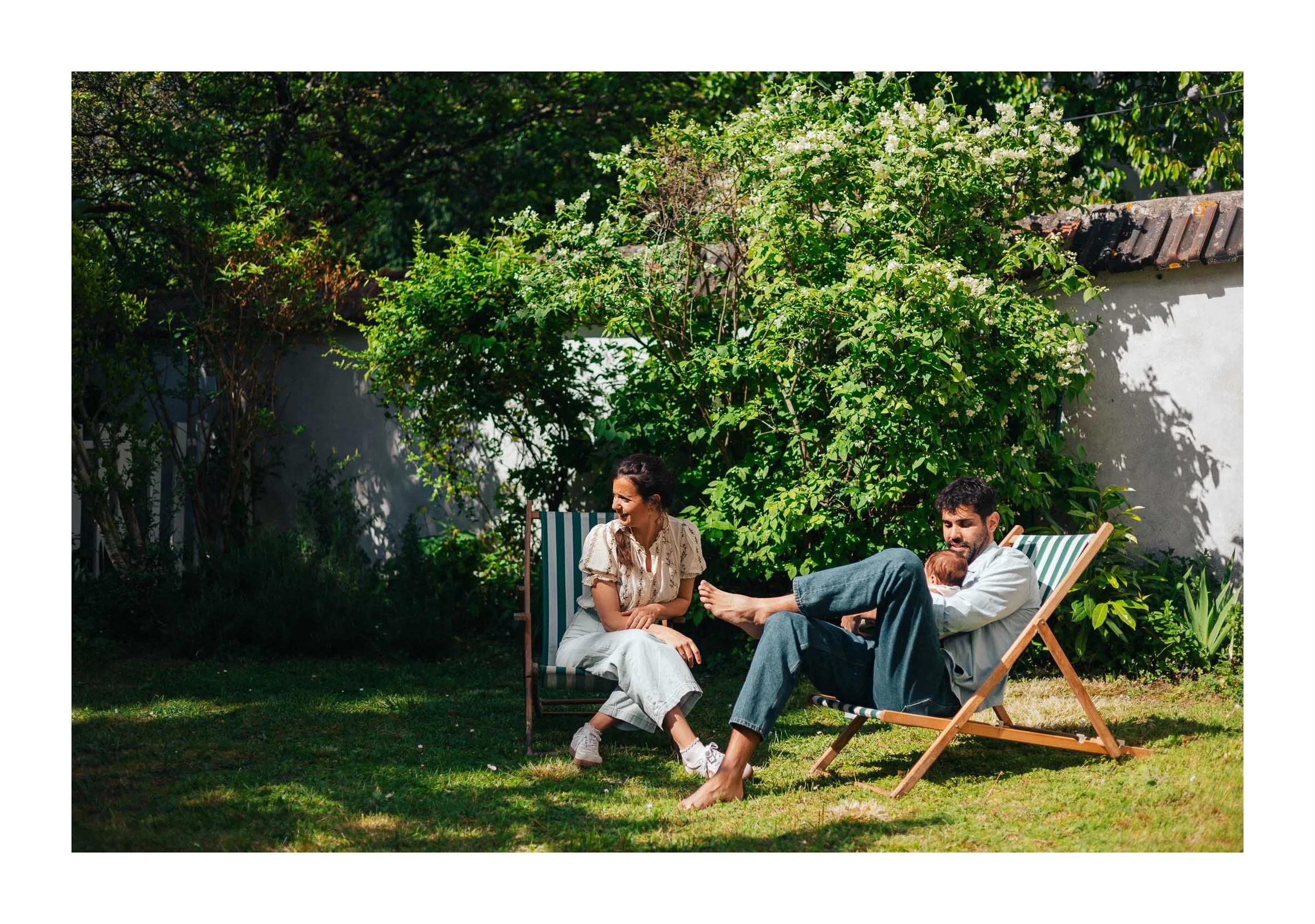 A group of three people sitting and relaxing outdoors in a garden, with lush greenery and flowering plants in the background.