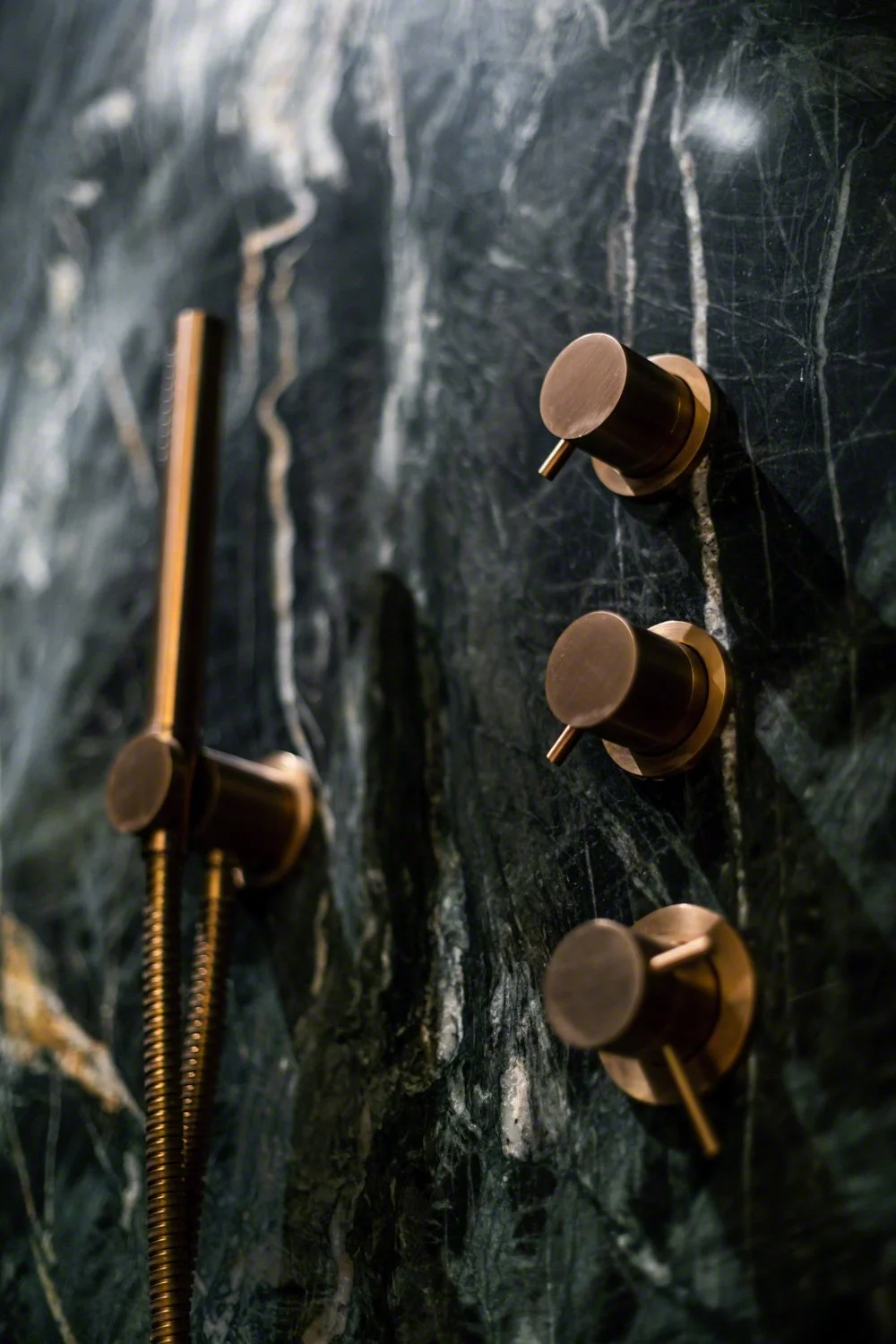 Close-up of three modern bronze shower knobs mounted on a black marble wall, with a brass shower hose and handle nearby.