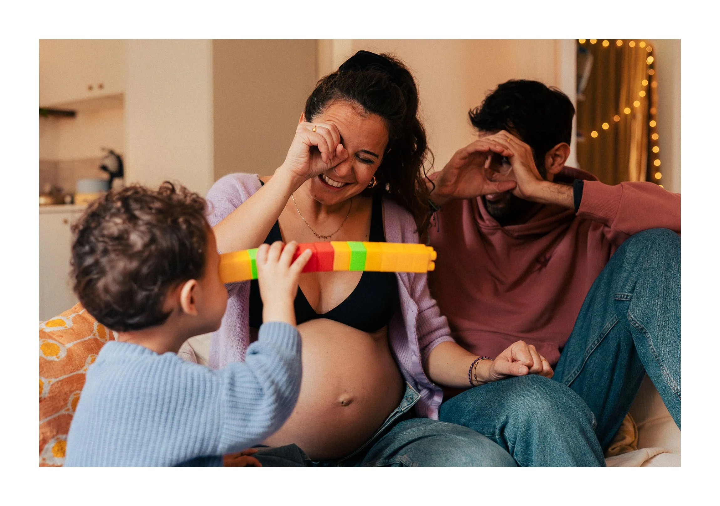 A pregnant woman with a big smile, sitting on a couch with her partner and a child, playing a game with colorful blocks, in a cozy living room with warm lighting.