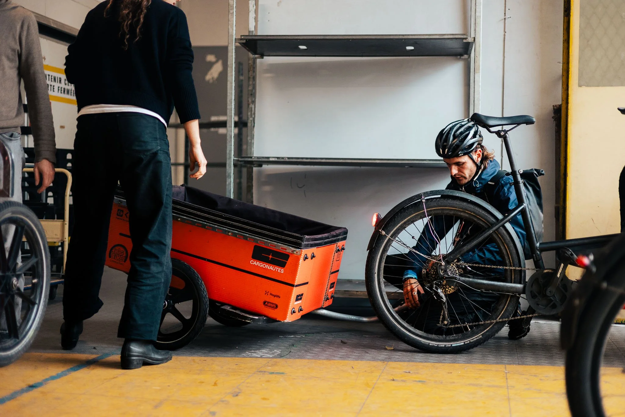 A person with a helmet working on a black bicycle with a cargo trailer attached, inside a storage or warehouse space.