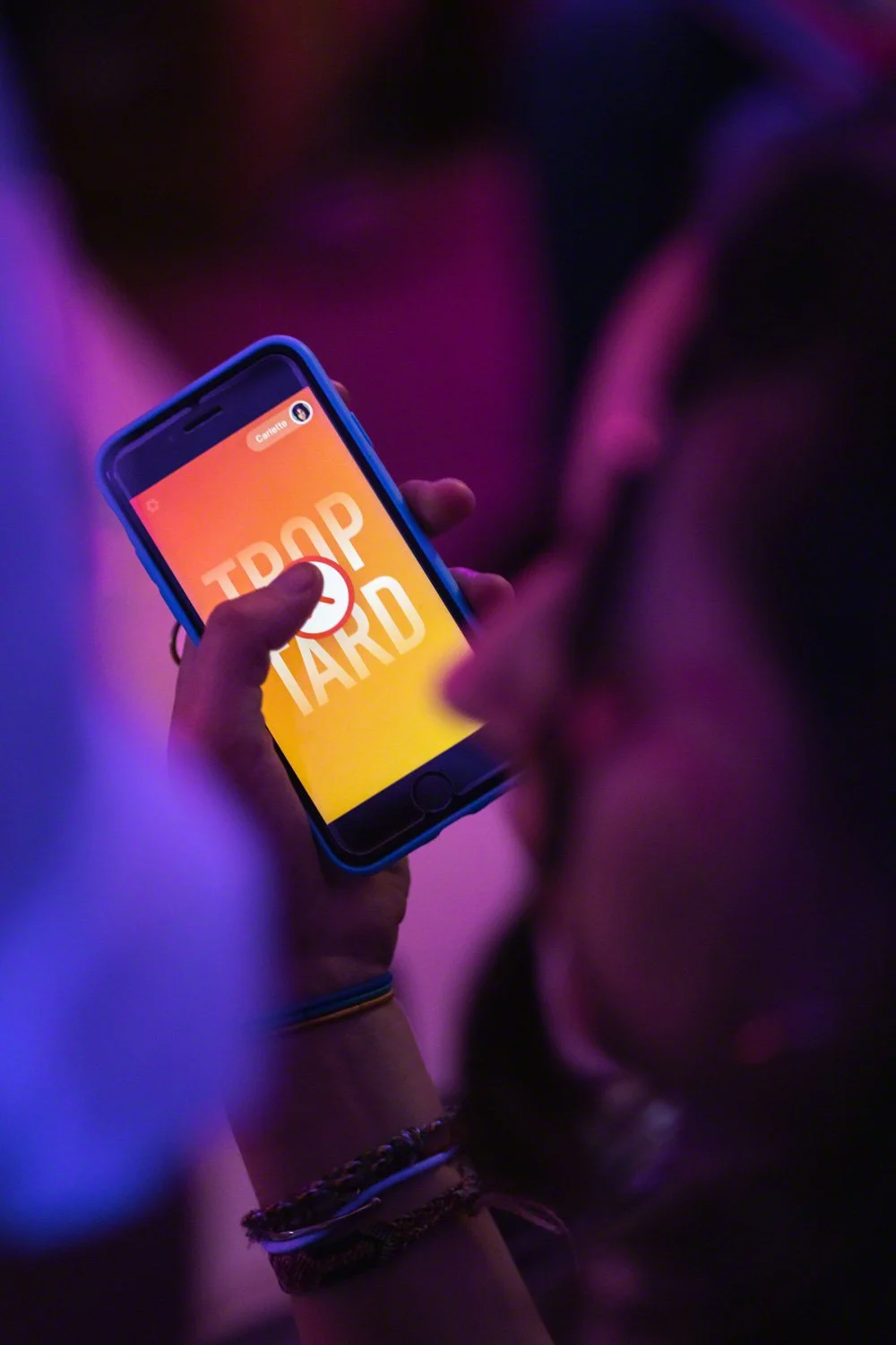 Person holding a smartphone displaying a colorful screen with the words "DROP TARD" in it, in a dark area with purple and blue lighting.