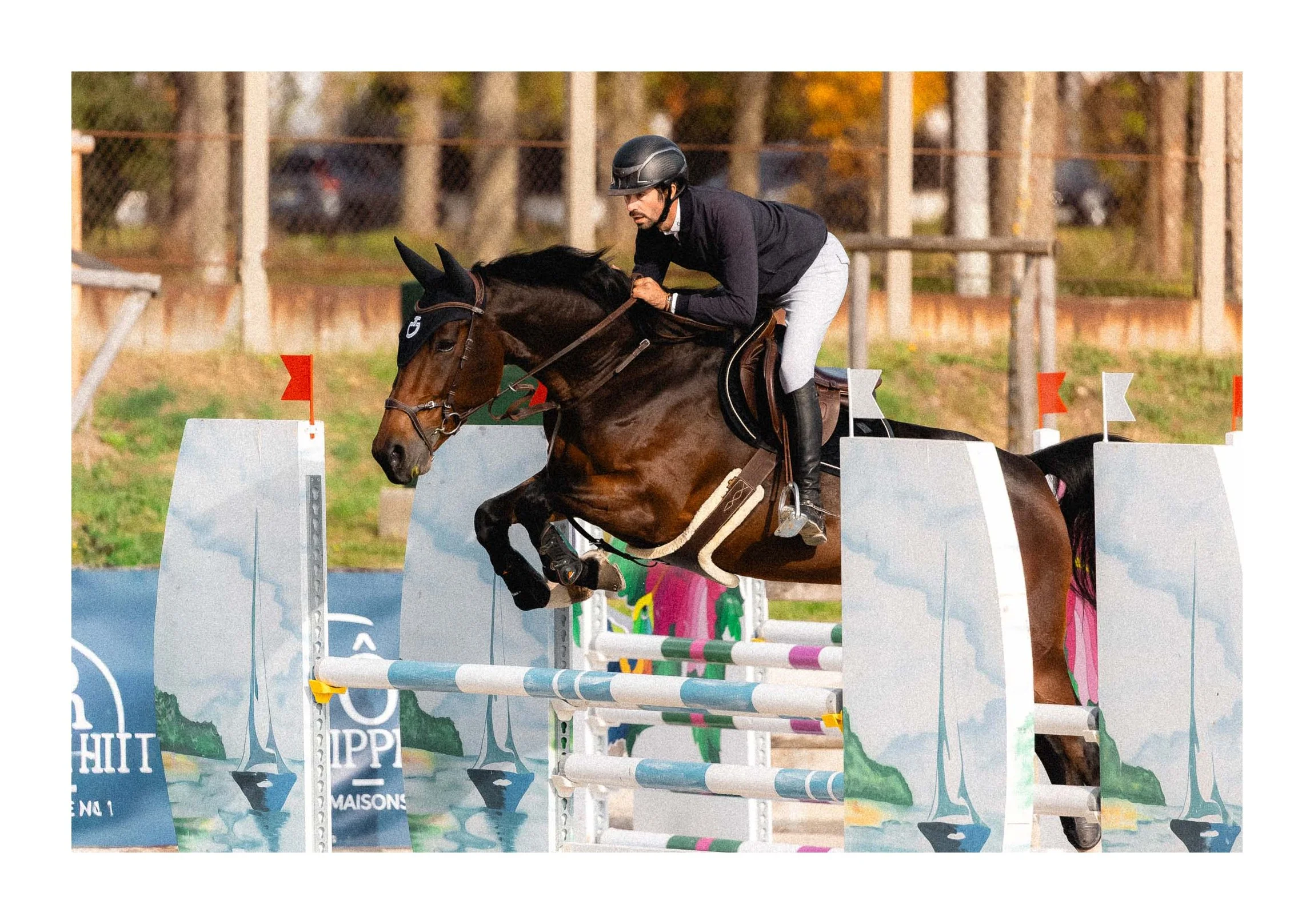 A male equestrian wearing a black helmet, black jacket, and white pants riding a brown horse and jumping over an obstacle in an outdoor horse jumping event.