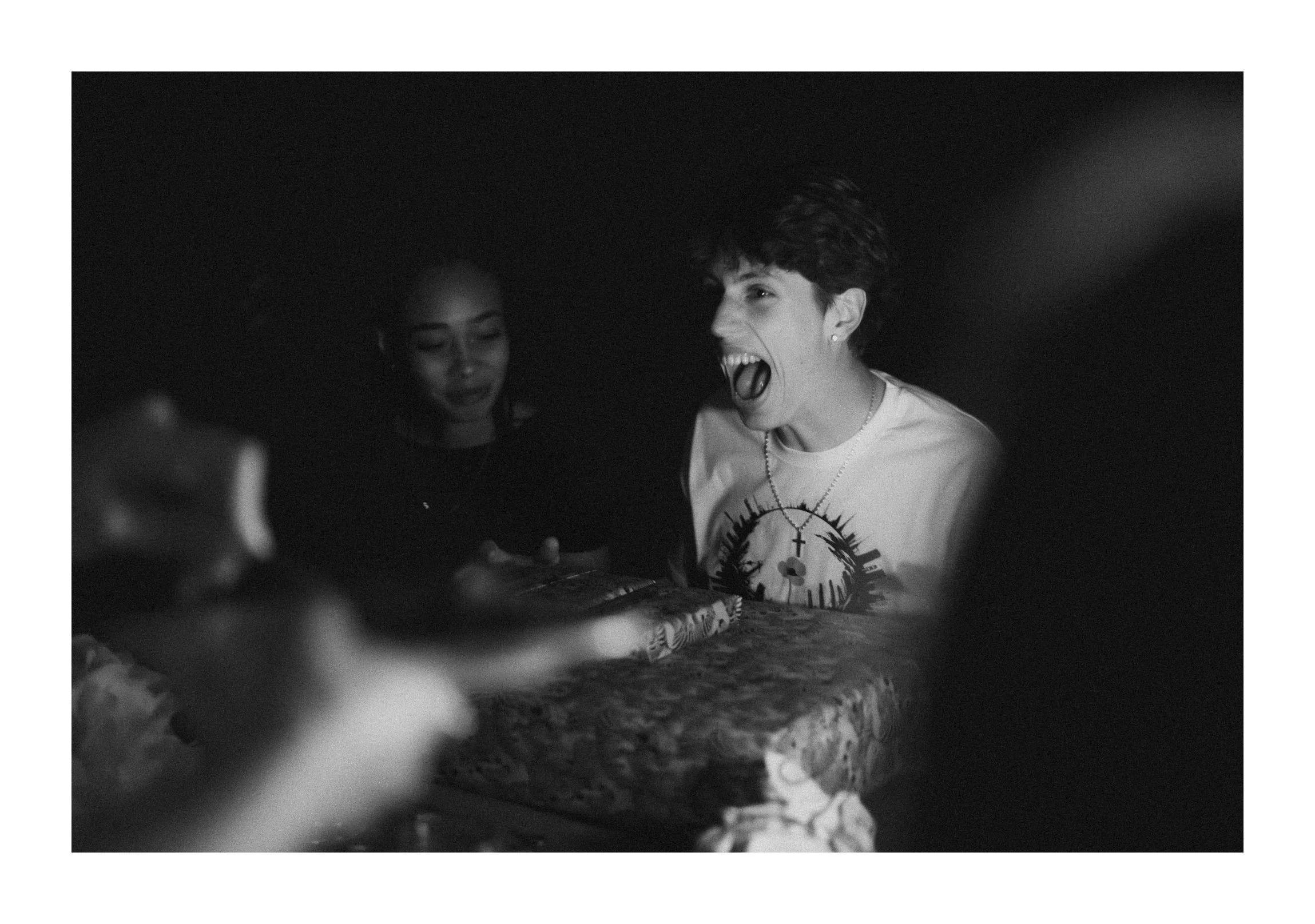 A person with short, curly dark hair is laughing or shouting while sitting at a table with a presence of a mailbox-shaped cake. In the background, there is a woman with straight hair looking on. The image is black and white.