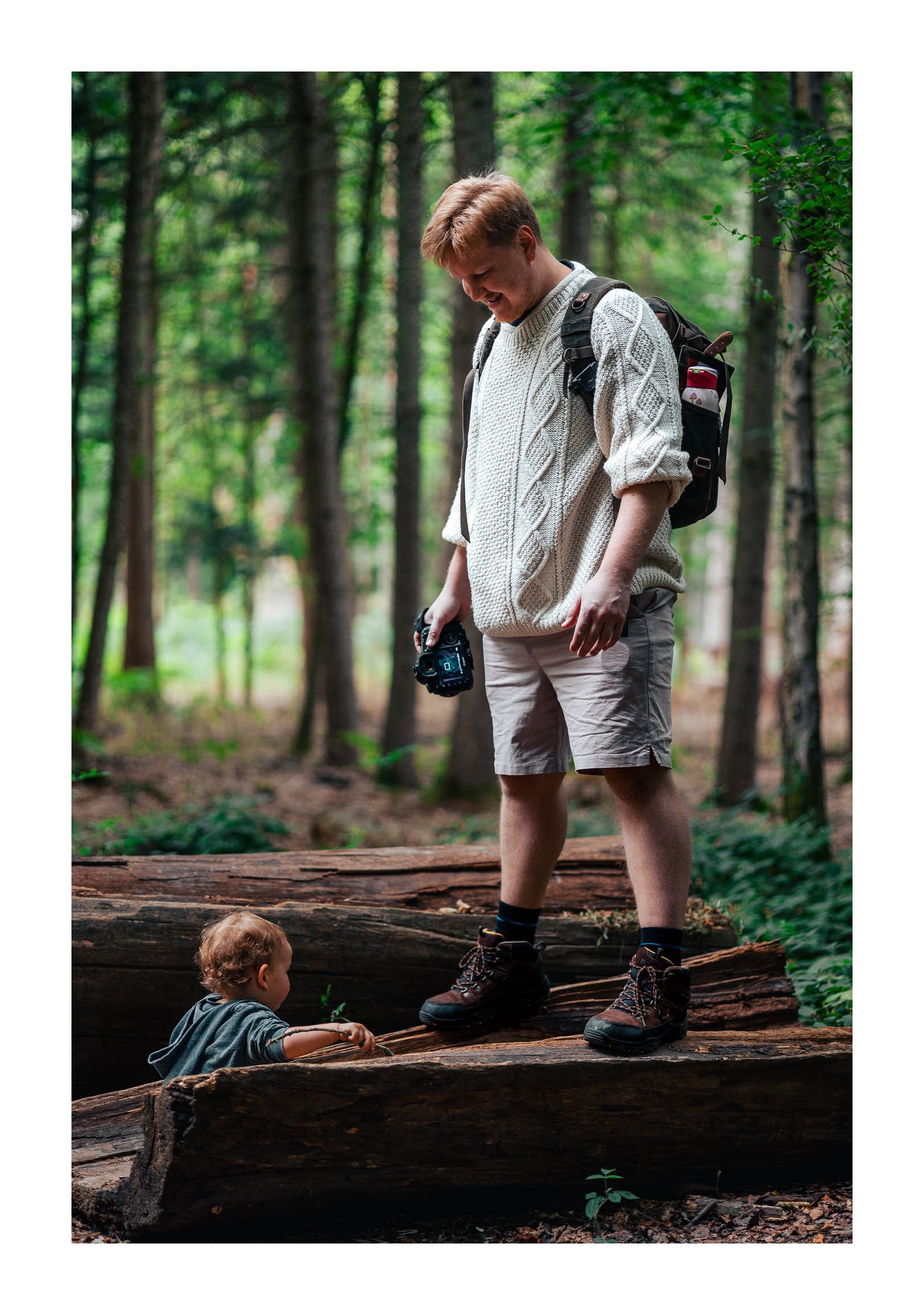 A man and a young child in a forest, with the man standing on a fallen log, holding a camera, wearing hiking boots, a backpack, and a light sweater. The child is sitting on the log, looking up at the man.