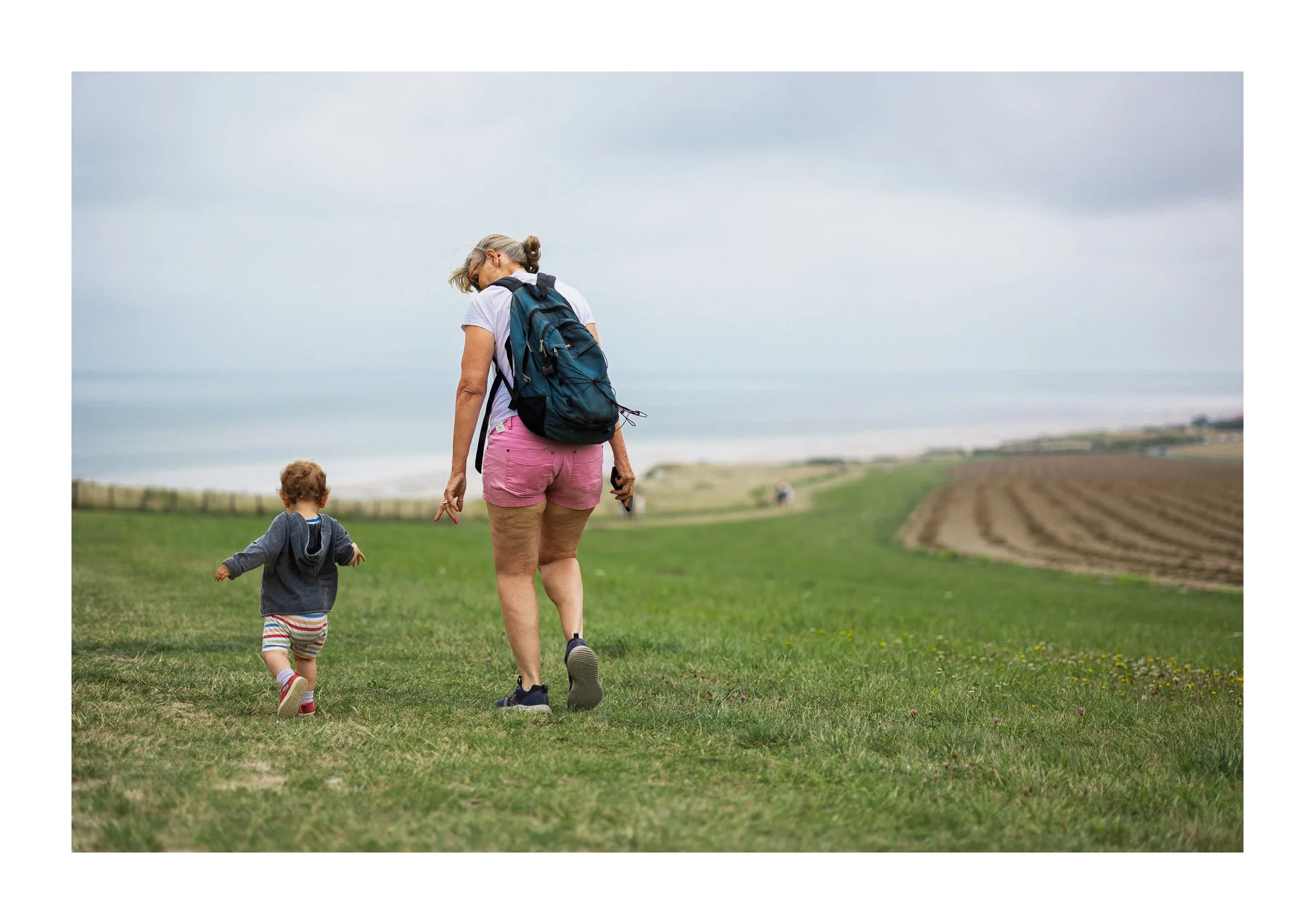 A woman and a young child walking on a grassy path near the coast, with fields and the ocean in the background on a cloudy day.