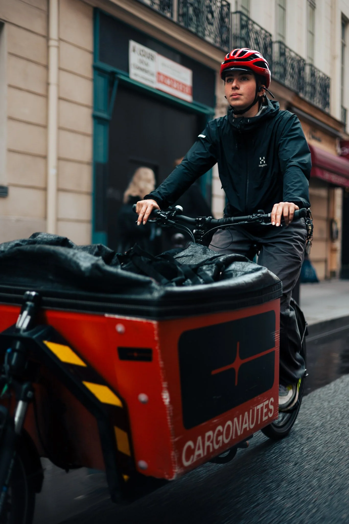 A woman wearing a red bicycle helmet and black jacket riding a cargo bicycle with a red and black cargo box labeled "Cargonautes" on a city street.