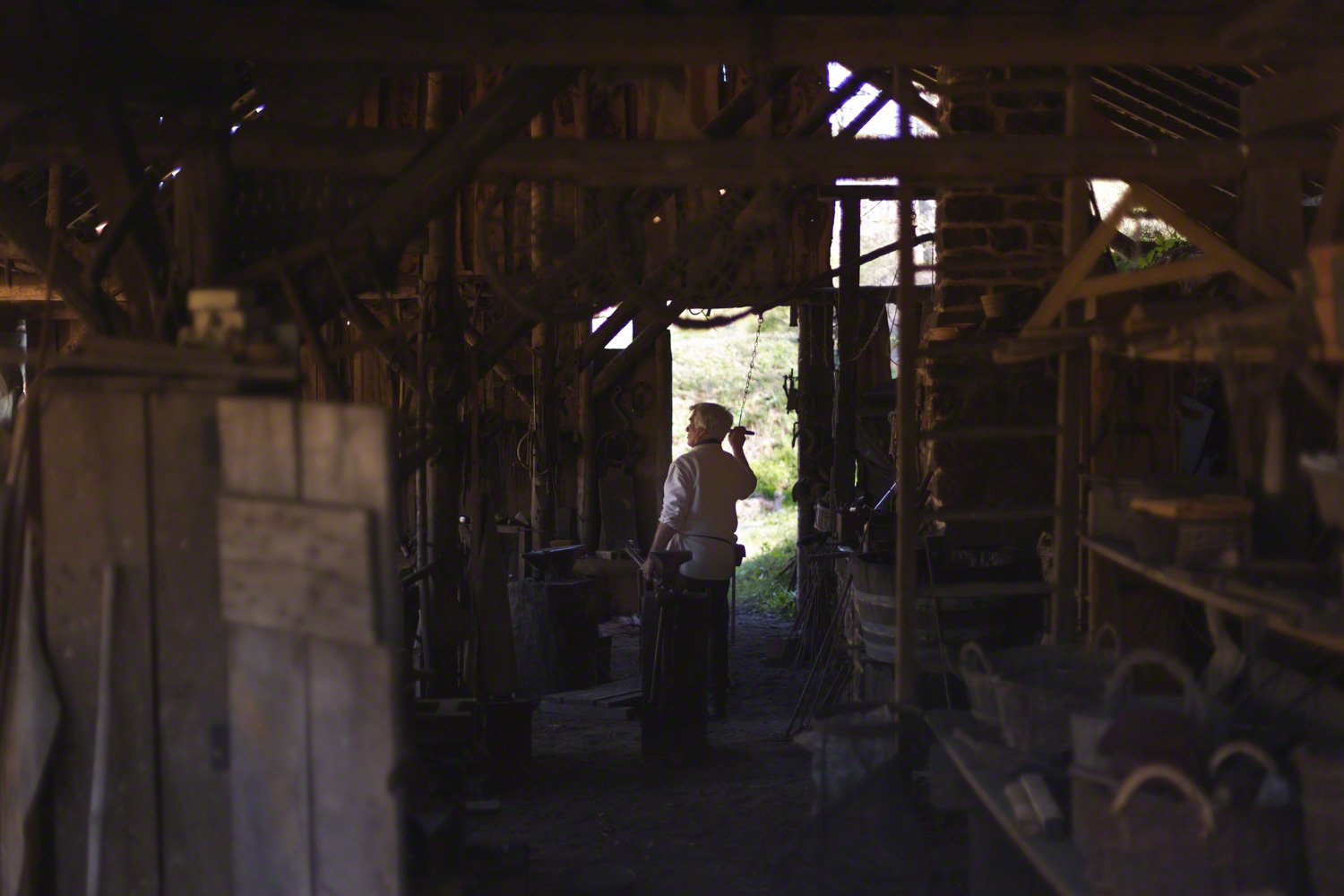 A person working in a rustic, wooden workshop with tools and equipment hanging on the walls, with light coming through an open doorway revealing a green outdoor landscape.