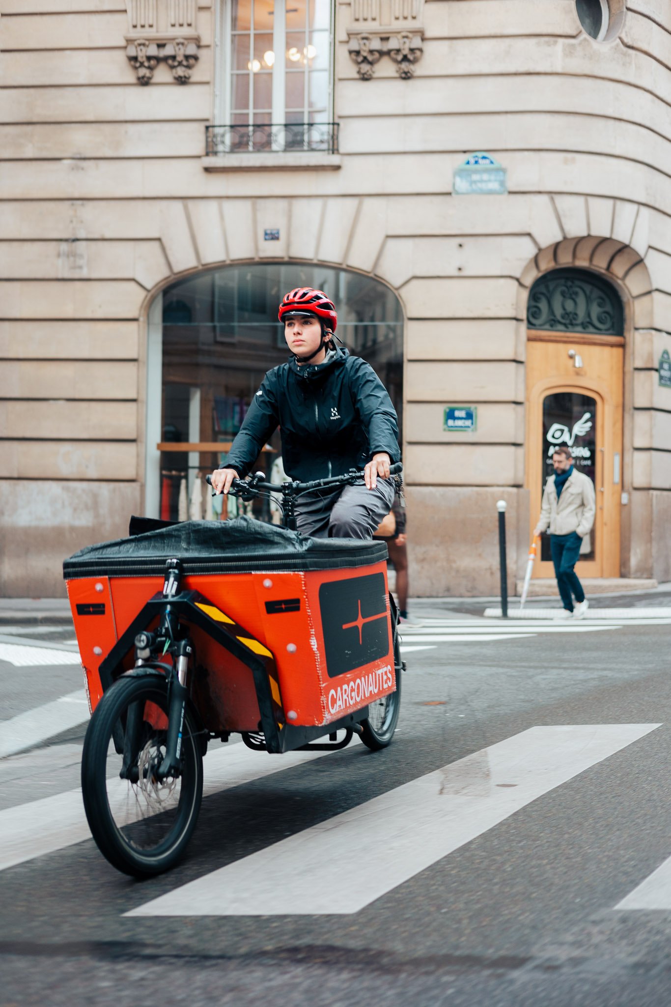 A cyclist wearing a red helmet rides a cargo bike with a large orange container labeled 'CARGONAUTES' across a city street with buildings and pedestrians in the background.