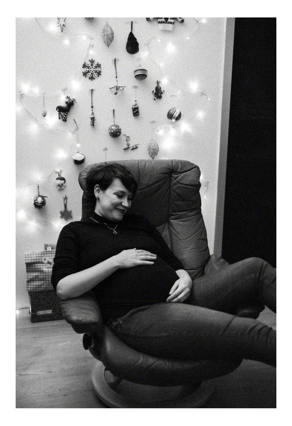 A pregnant woman sitting in a recliner, smiling and resting her hand on her belly, in front of a decorated wall with Christmas ornaments and lights.