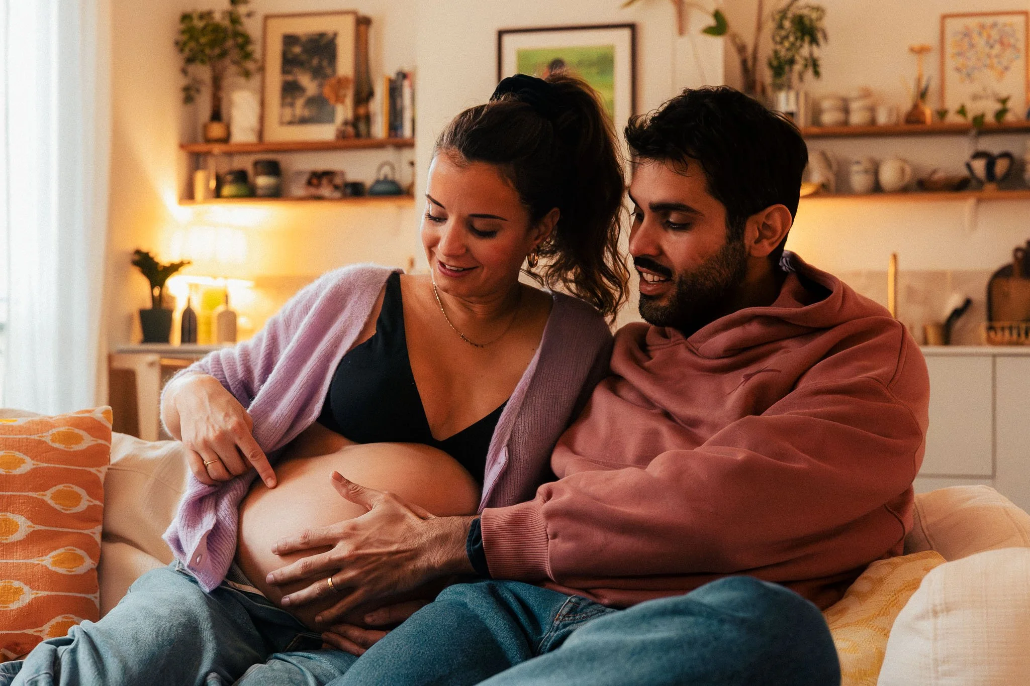 A pregnant woman and a man sitting together on a couch, touching her baby bump and smiling in a cozy, well-lit living room with shelves and framed artwork in the background.
