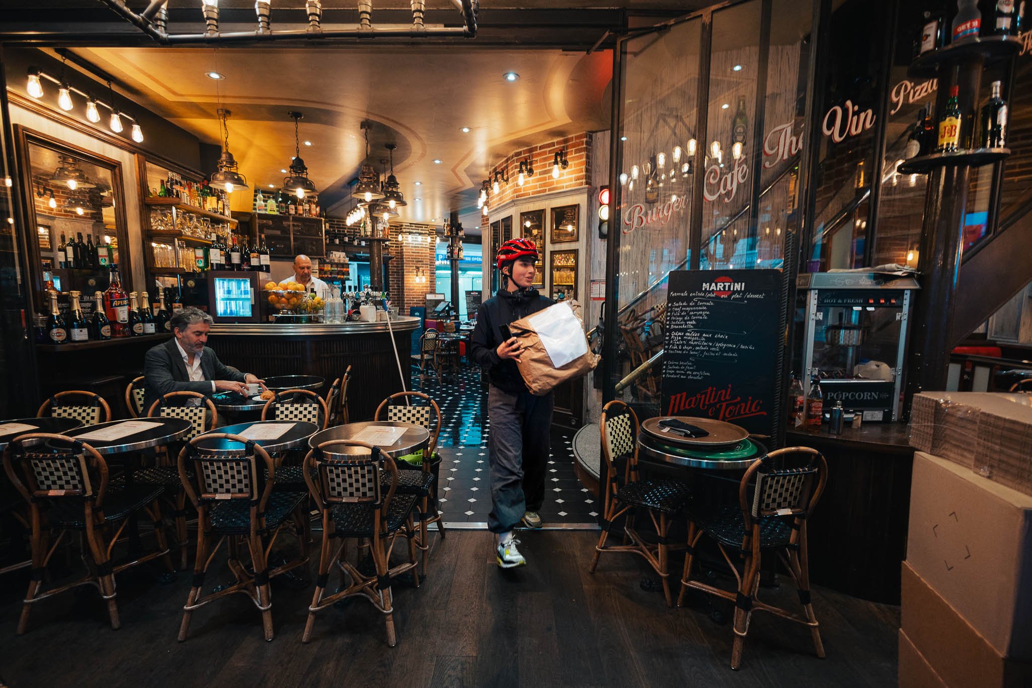 A person wearing a red helmet and dark clothing is carrying a paper bag inside a cozy, dimly lit restaurant or cafe. The interior features wooden chairs with checkered seats, a black and white tiled floor, and a bar area with bottles and glasses behi