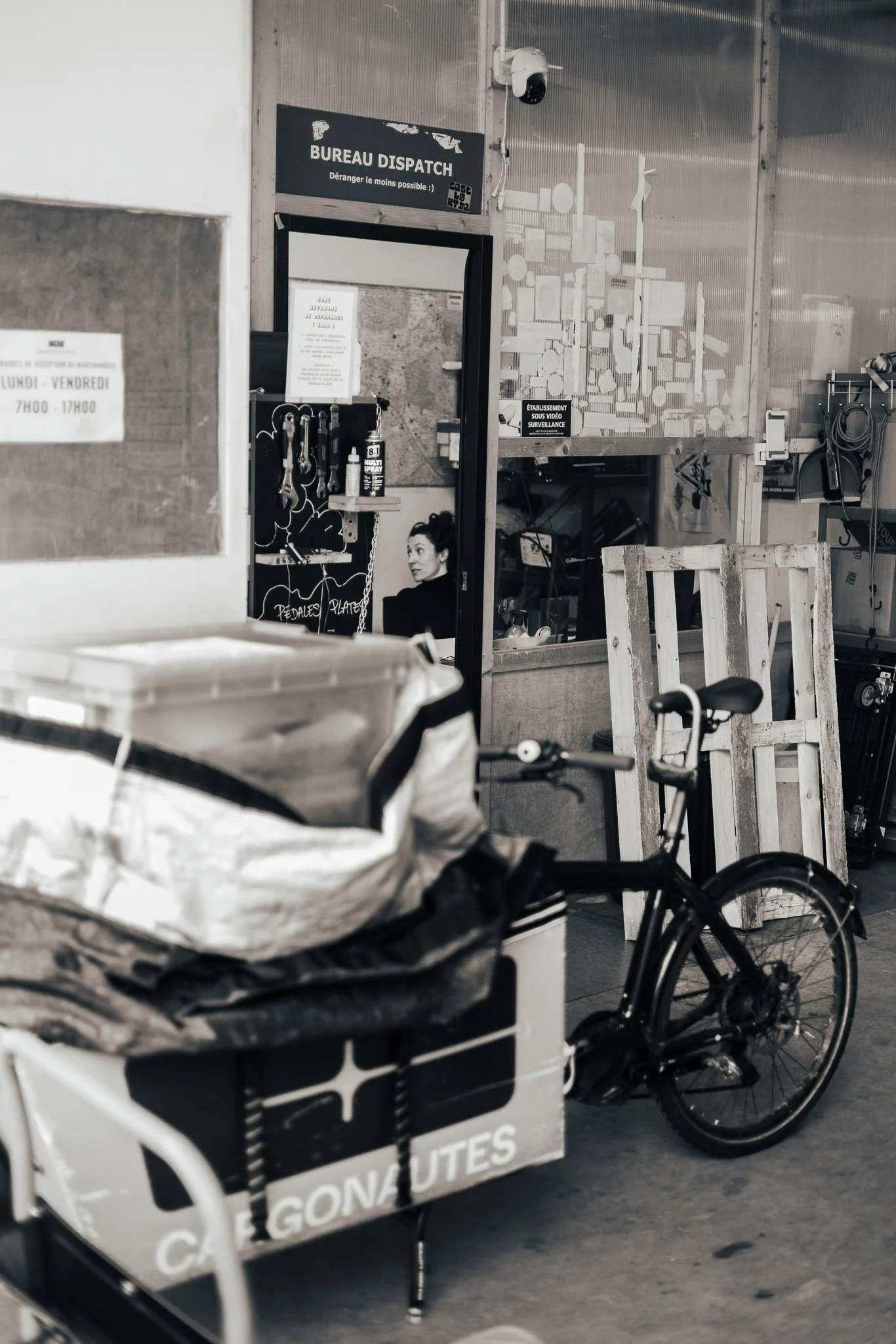 A black bicycle is parked next to a cart with a cardboard box reading 'CAFE GONAUTES' in an indoor space. A woman with dark hair tied up is sitting inside a room beyond a doorway, looking towards the camera. The surroundings include various equipment