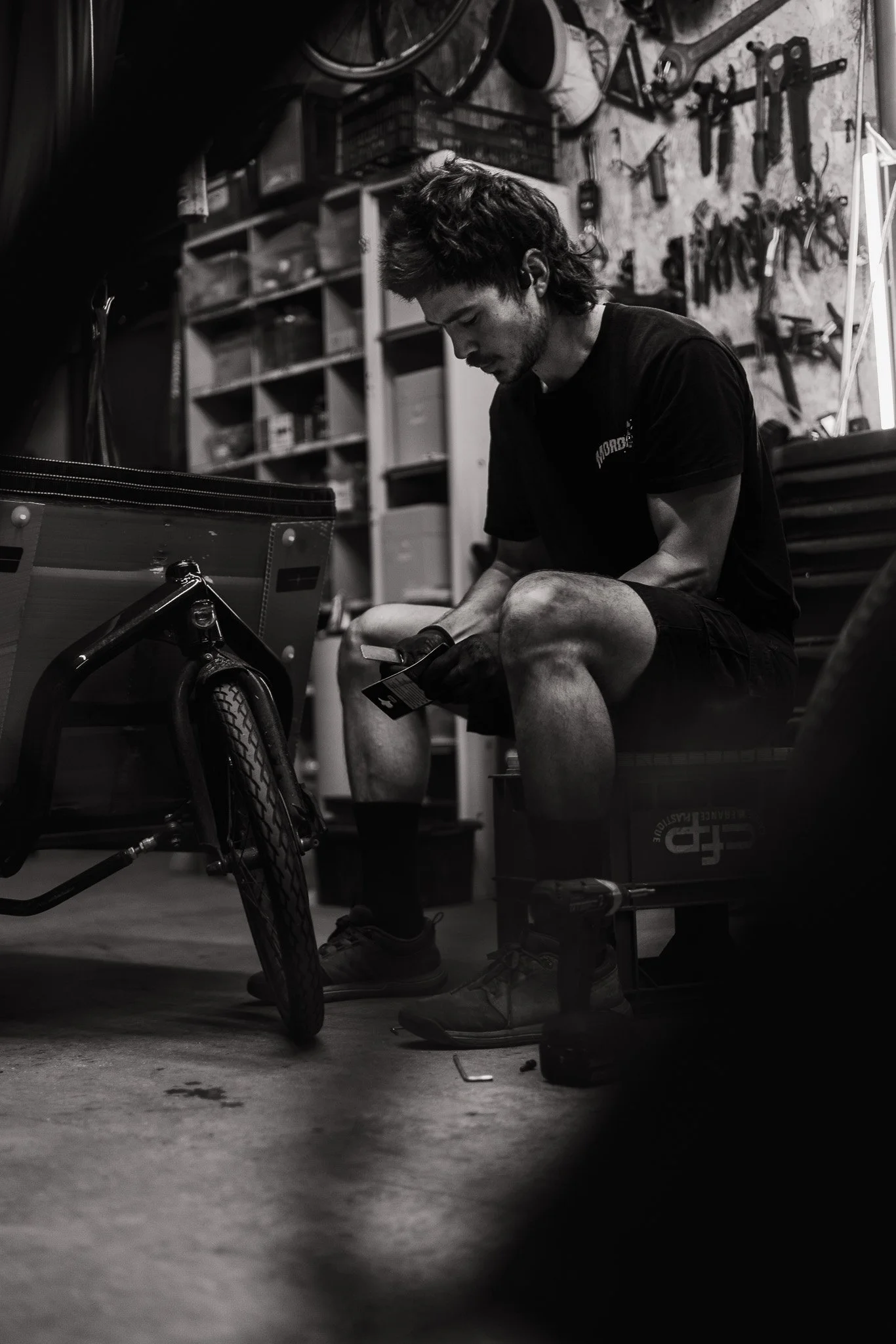 A man sitting on a bench in a workshop, looking at his phone with tools and equipment hanging on the wall behind him.