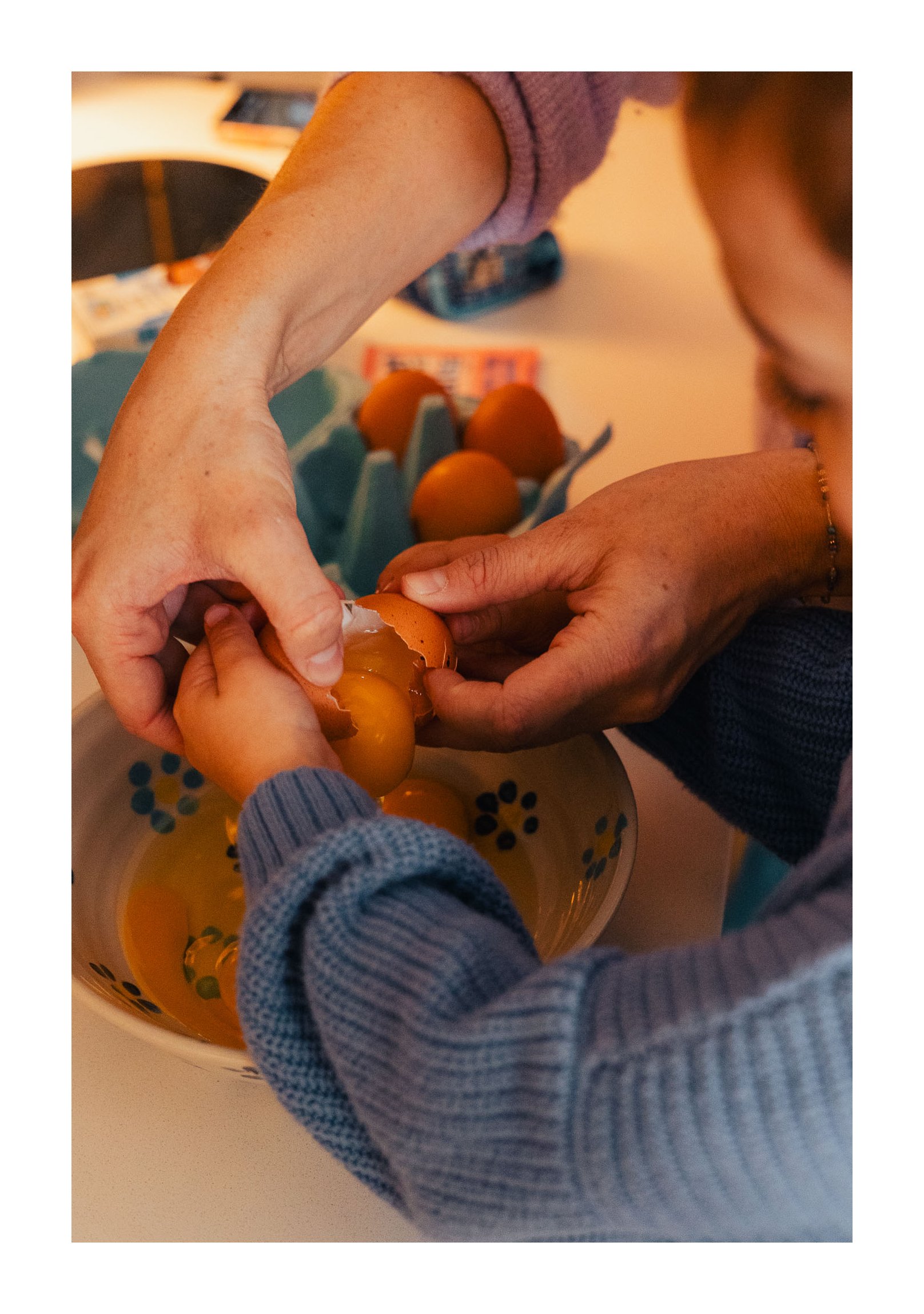 Two people cracking eggs into a bowl, with more eggs and kitchen items visible in the background.