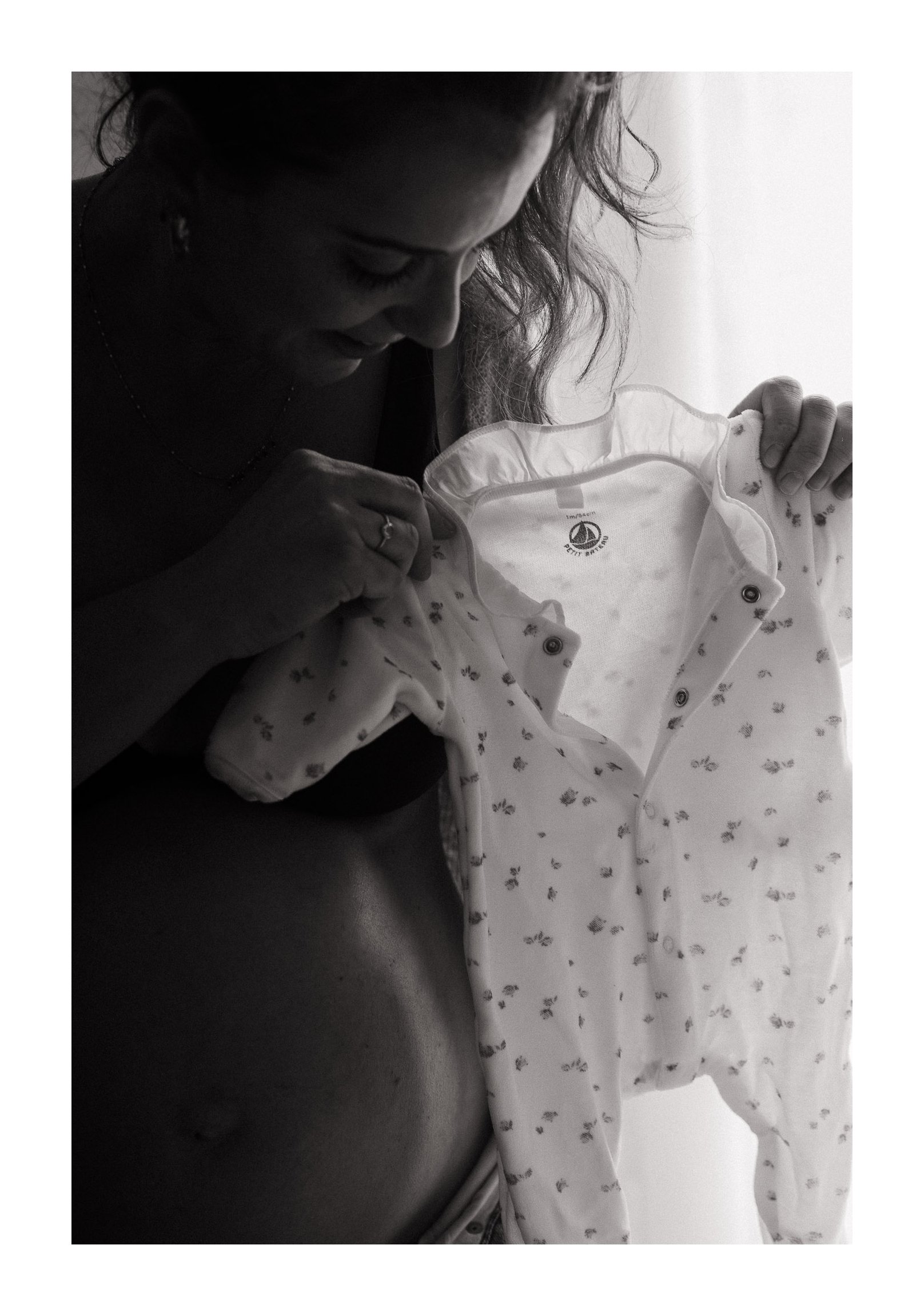 A woman is holding a light-colored baby outfit with tiny dark patterns, looking at it lovingly.