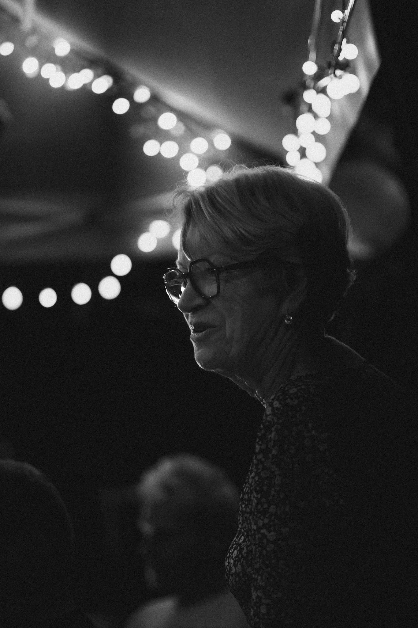 A black and white photo of an elderly woman with glasses, smiling at a social event, with string lights in the background.