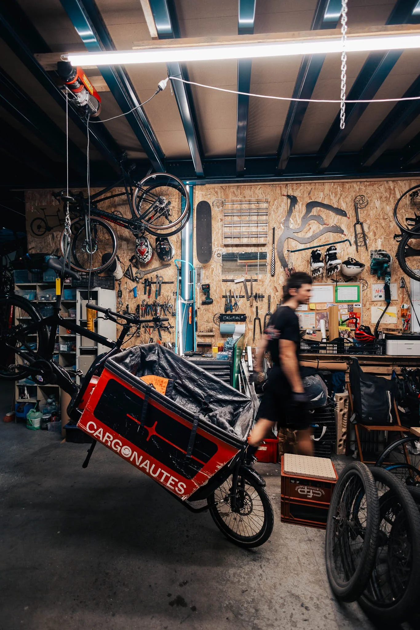 A workshop with walls covered in tools, bikes, and skateboards. A person is walking past a bike with a cargo box labeled 'Cargonautes' filled with items. The area is cluttered with bicycle parts and storage bins.