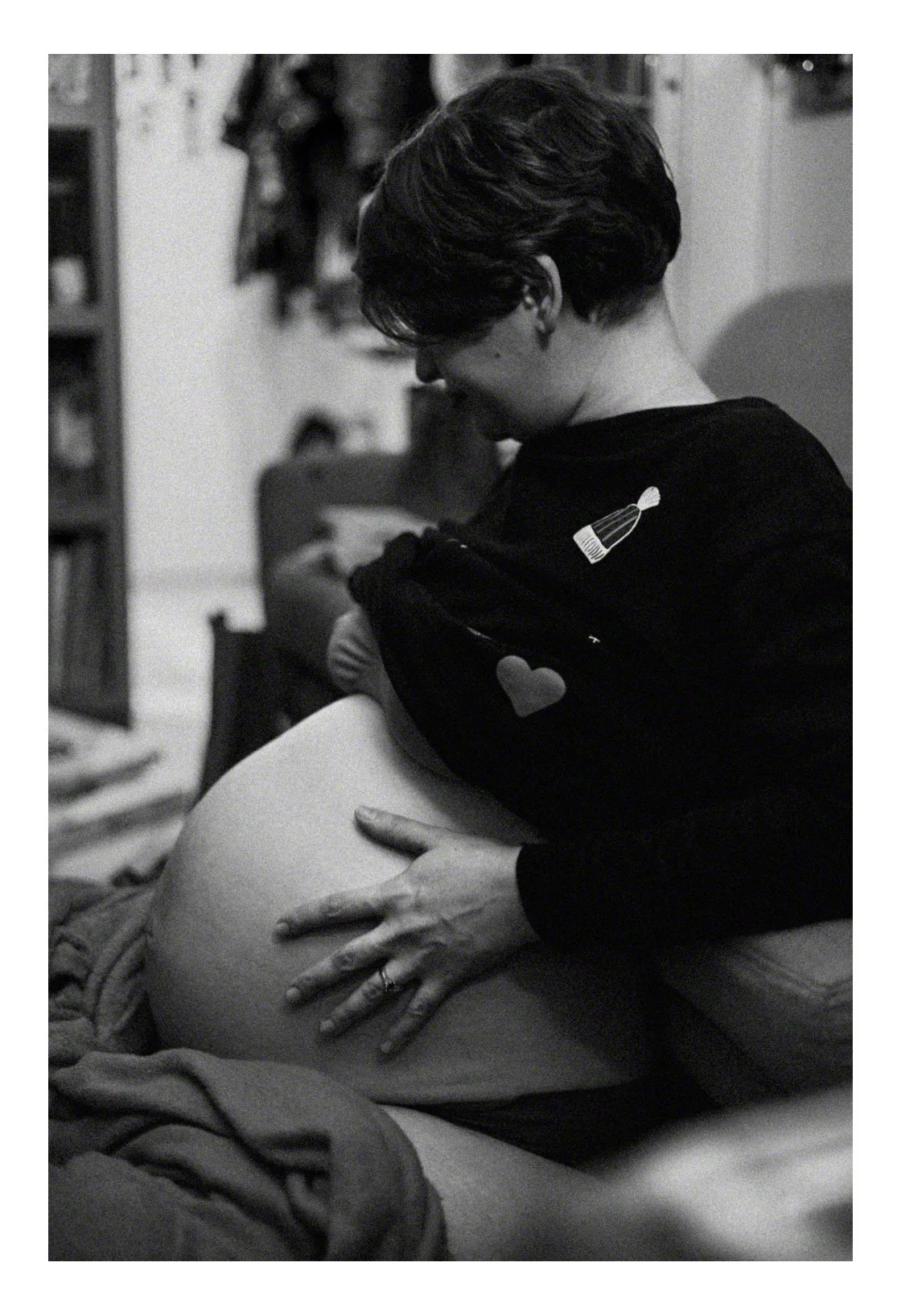 A woman with short hair and glasses, wearing a dark sweater with a patch of a bottle and a heart, sitting on a bed and holding a pregnant belly with one hand while lovingly pressing her face against it.