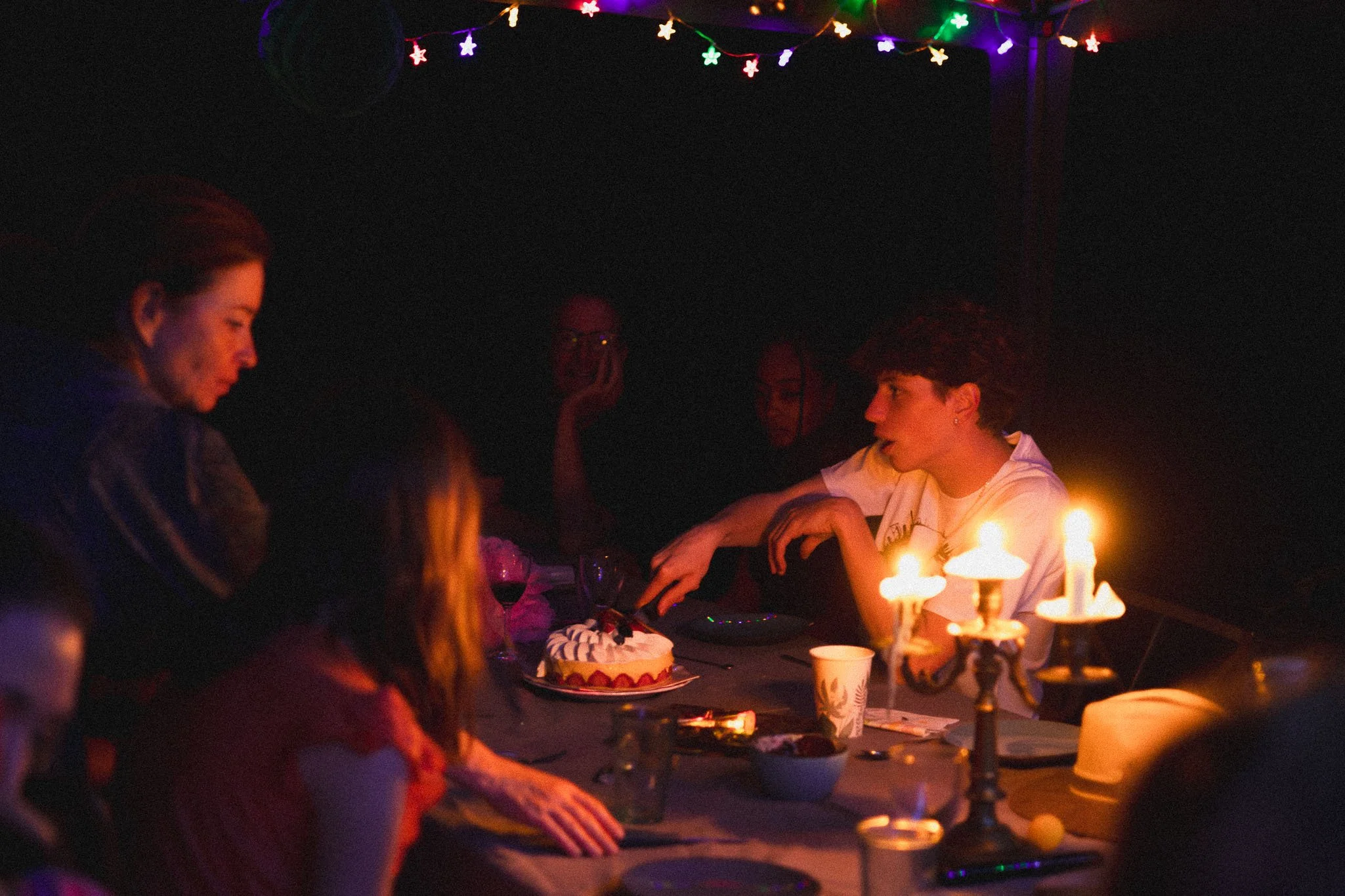 People celebrating a birthday at night with a cake featuring lit candles, under colorful string lights.