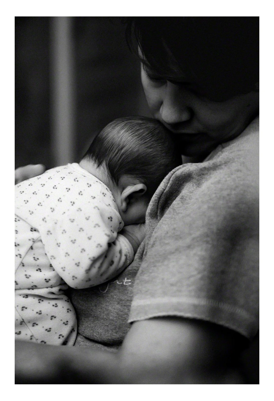 A black and white photograph of a woman holding and caring for a sleeping baby close to her chest, with the woman's face gently near the baby's head.