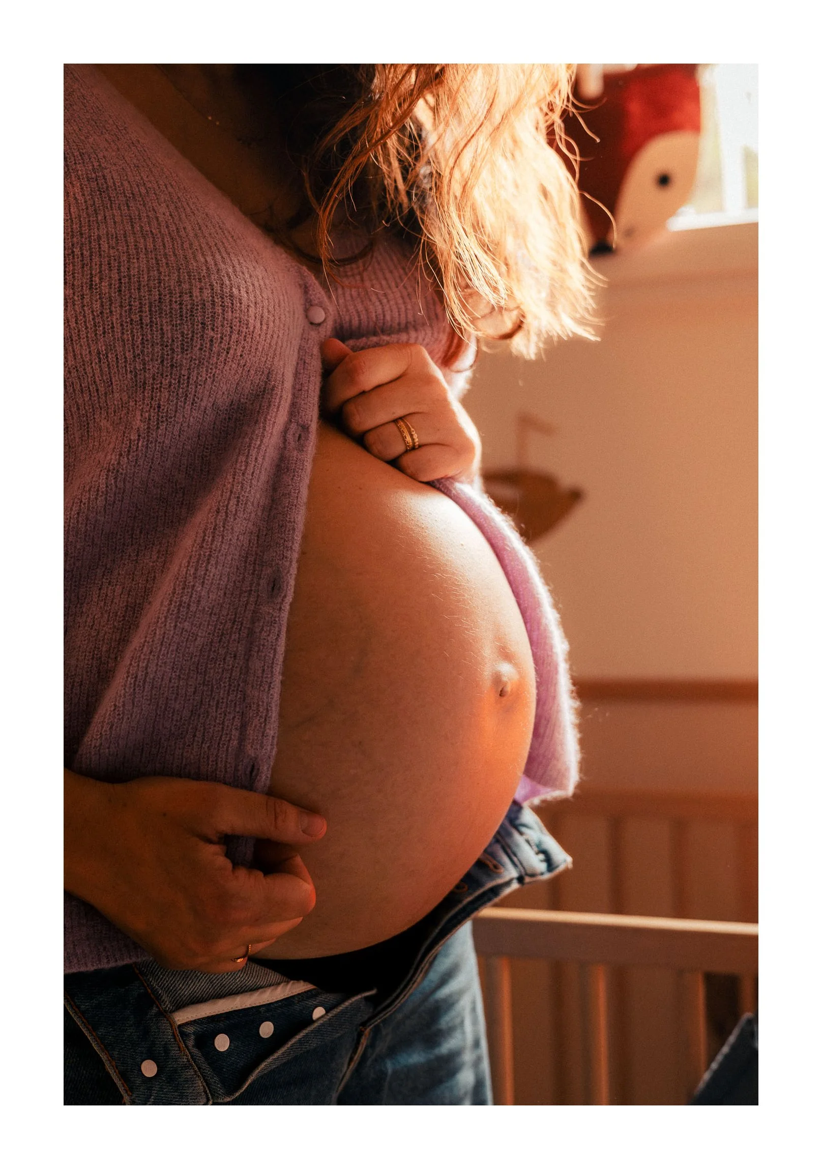 A pregnant woman gently touching her belly, wearing a purple sweater and jeans, with sunlight illuminating her hair.