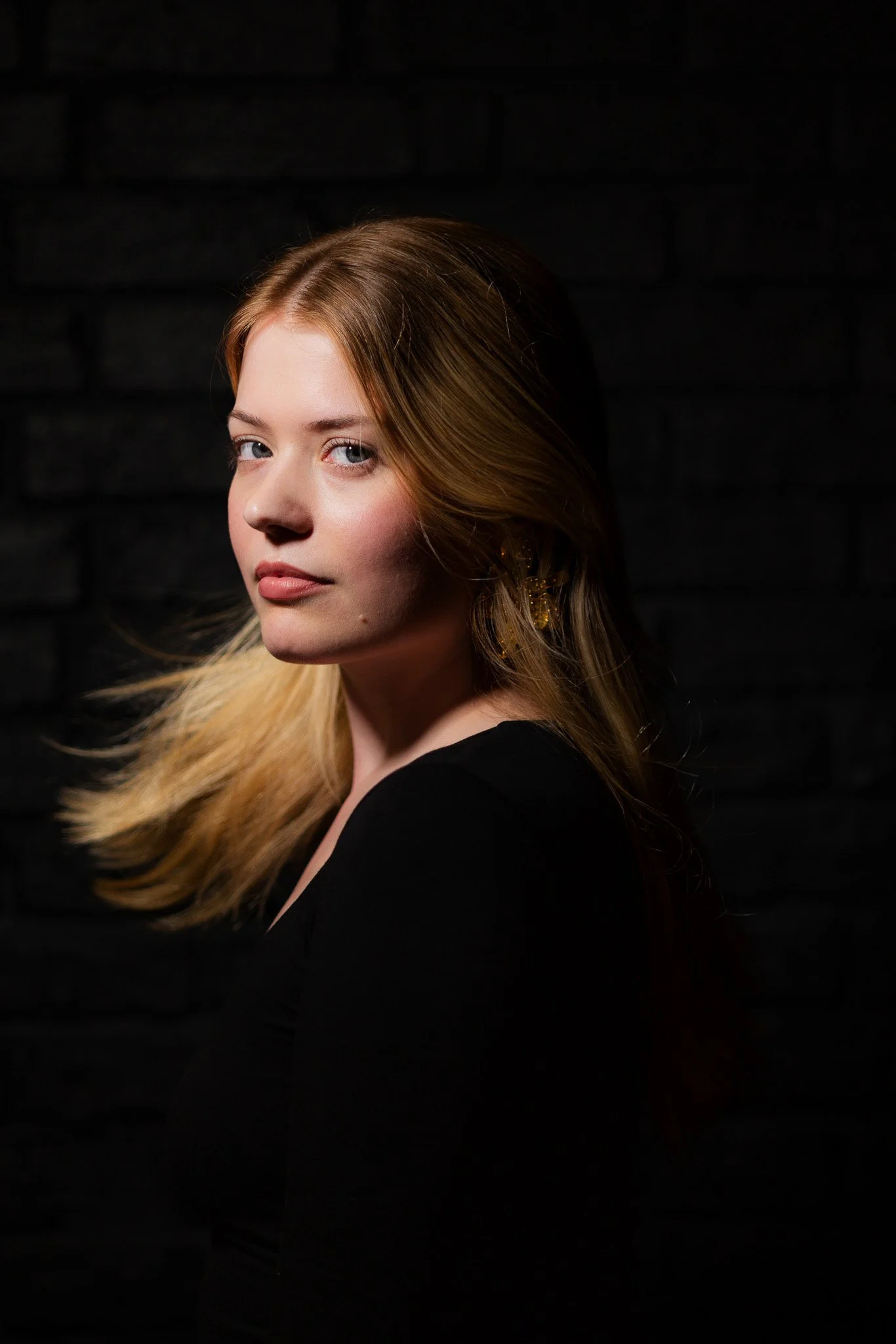 Portrait of a woman with blonde hair, looking at the camera against a black brick wall background.