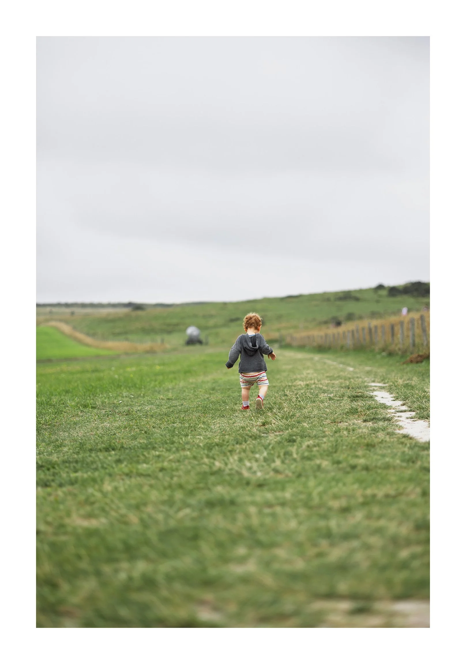 A young child with curly hair running on a grassy field under an overcast sky with hills and a fence in the background.