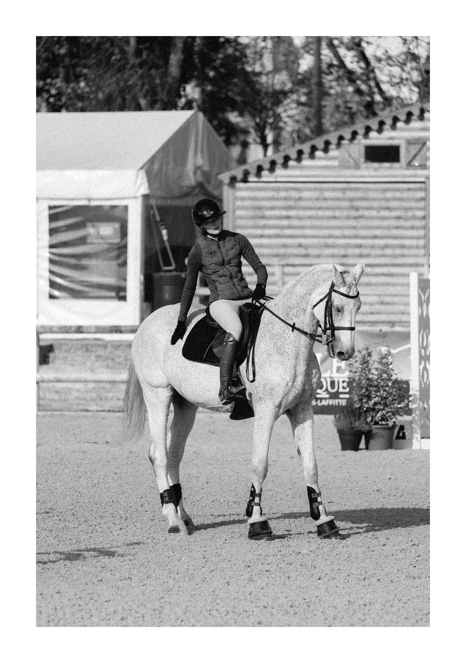 A person riding a white horse at an equestrian event in an outdoor riding arena, black and white photo.