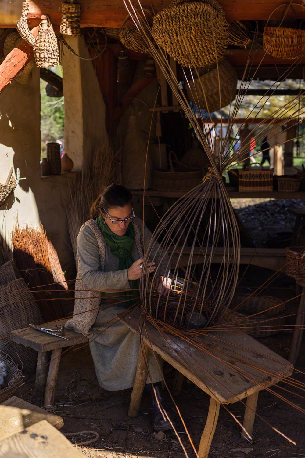 A woman working on a large, intricate wire sculpture inside a rustic workshop. The workshop has wooden shelves with woven baskets and other craft items, and sunlight filters through a small window.